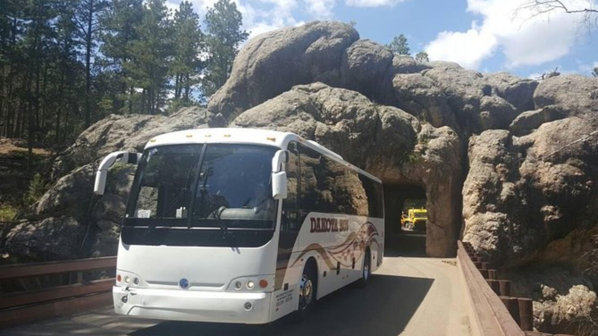 A large white tour bus exits a narrow tunnel carved through rugged rock formations, surrounded by pine trees under a partly cloudy sky.