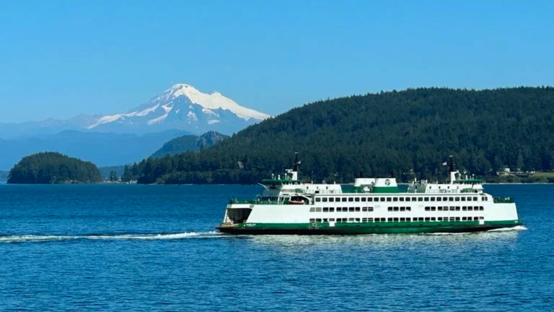 A green and white ferry sails on a blue ocean, with a forested island and a snow-capped mountain in the background under a clear blue sky.