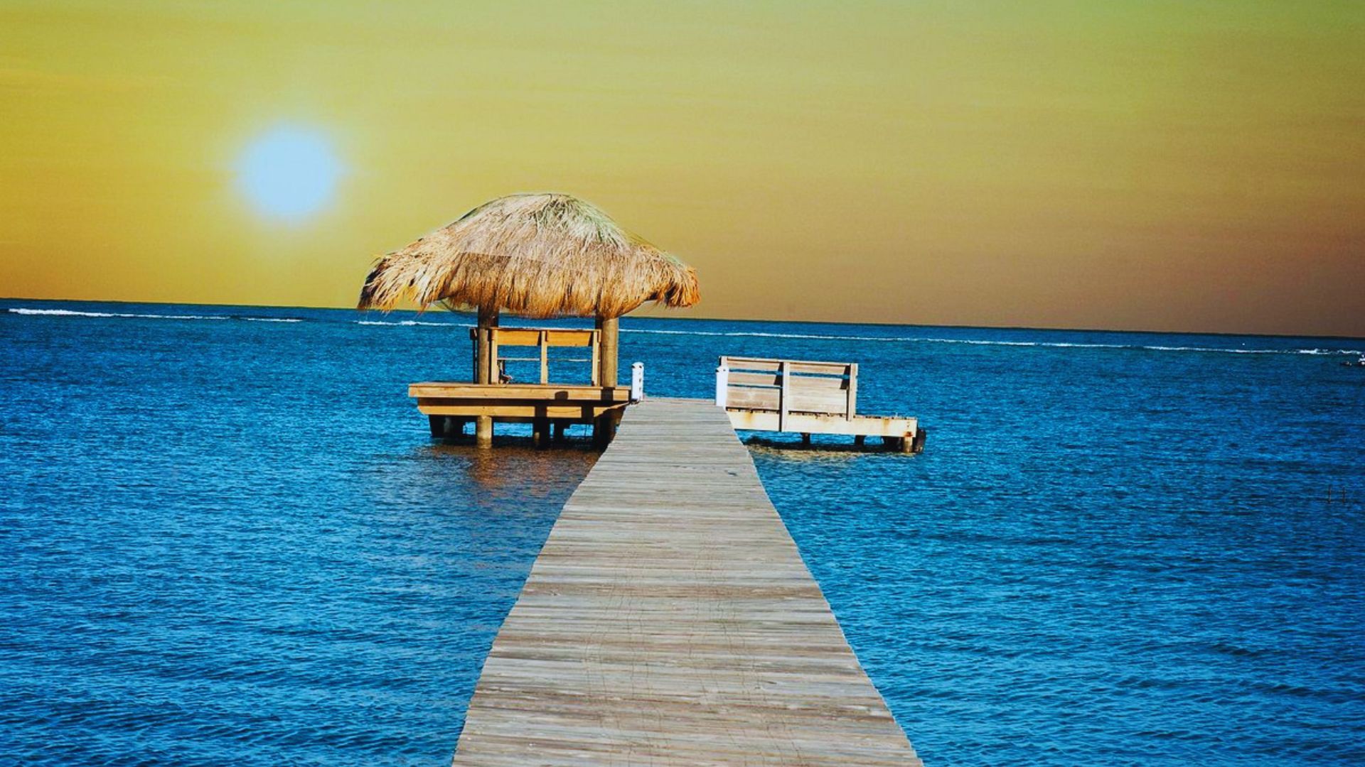A wooden pier leads to a thatched hut over calm blue ocean waters under a setting sun, conveying a peaceful, tropical sunset ambiance.