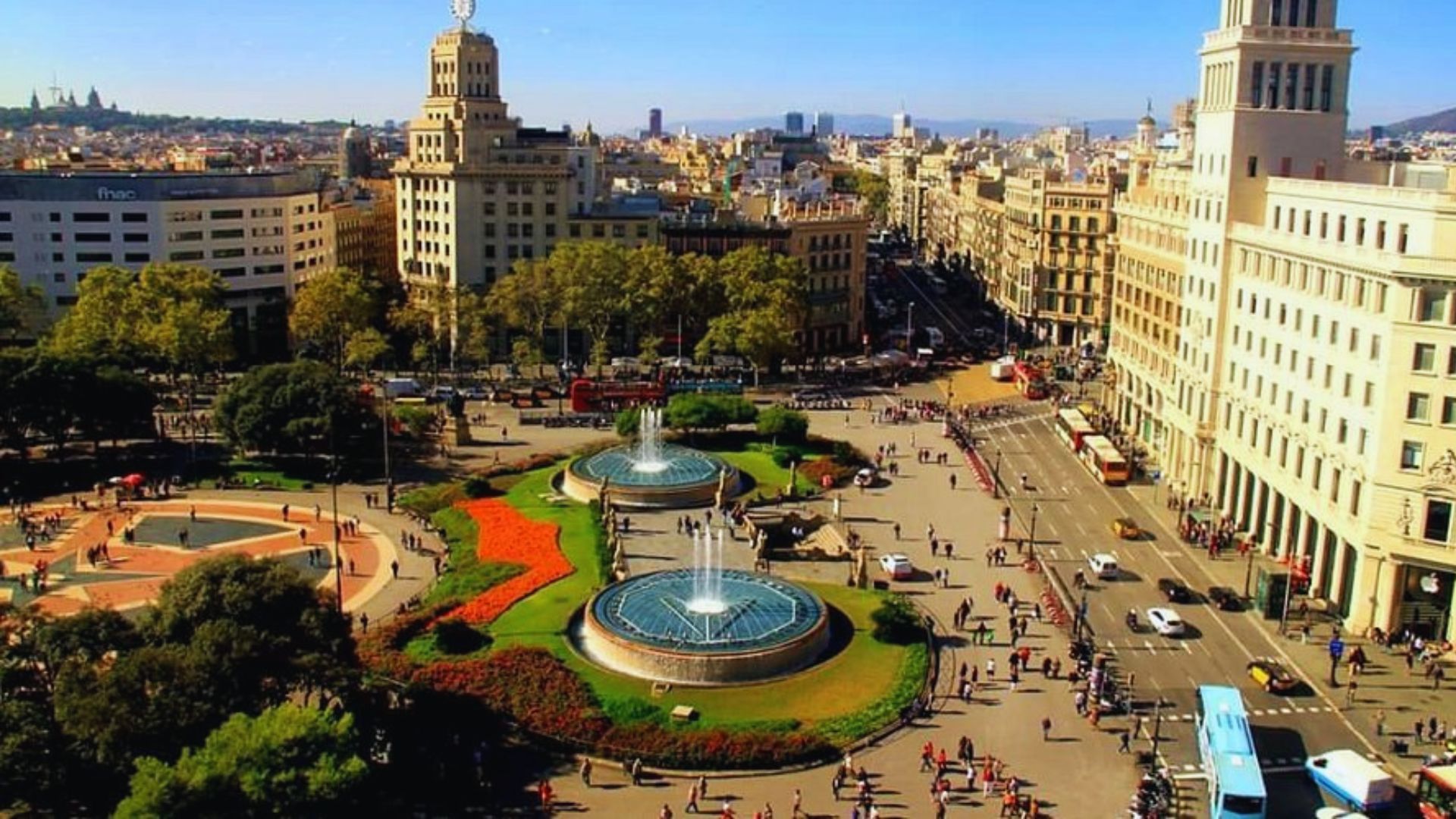 A bustling city square on a sunny day with two large fountains. Surrounded by historic buildings and trees, people walk and gather, creating a lively atmosphere.