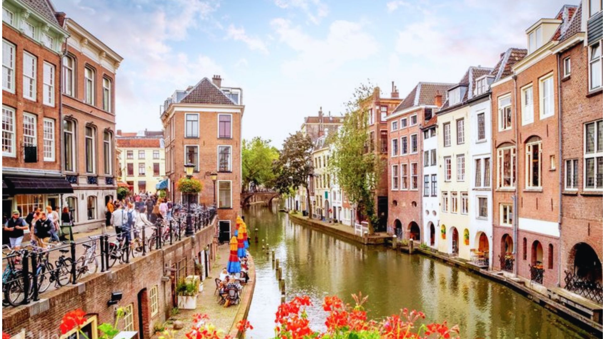 A picturesque canal in a European city lined with charming, colorful buildings. People stroll along the walkway beside red flowers. Bright, clear day.