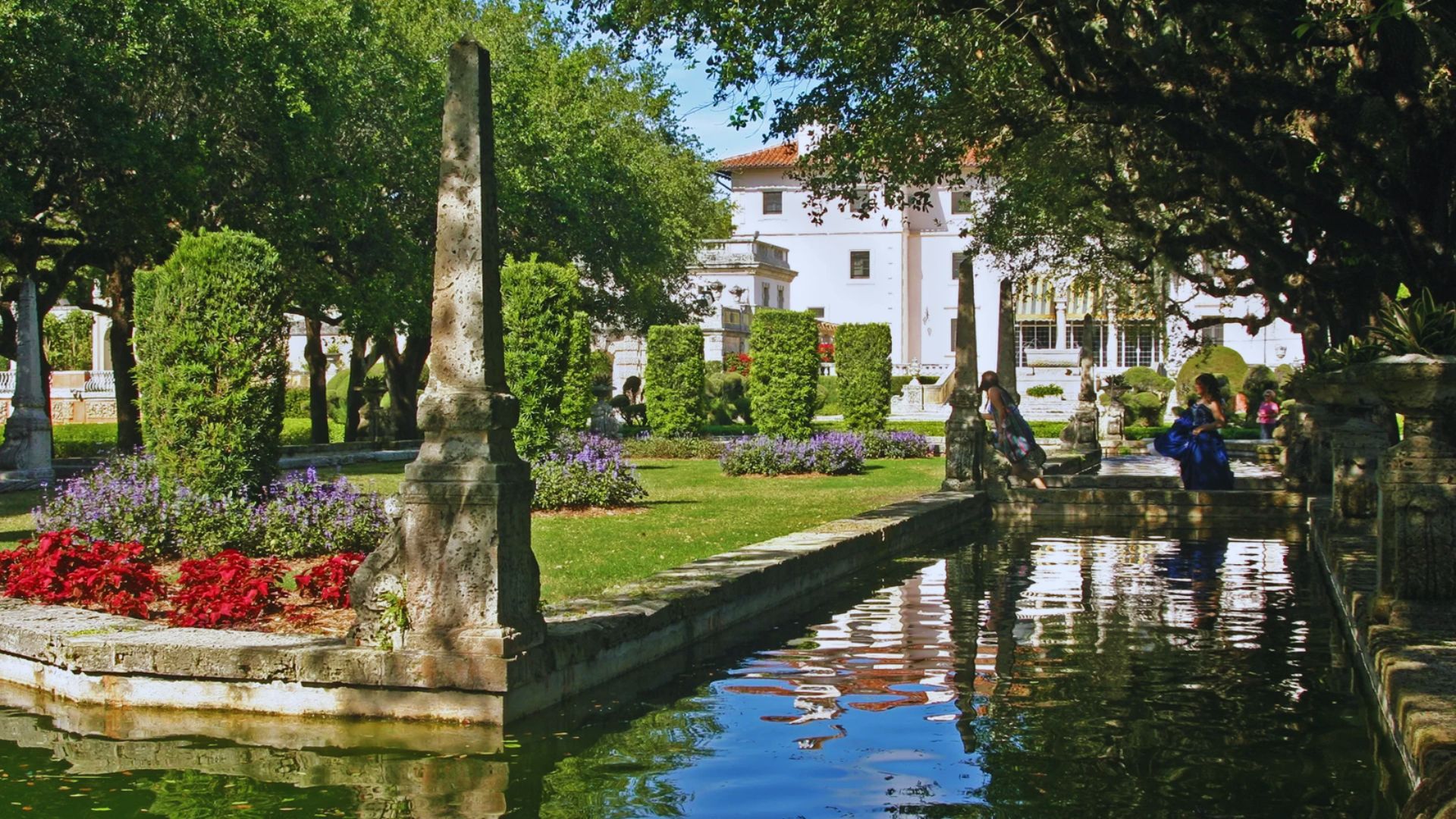 Lush garden with manicured hedges, a reflective pond, and blooming red and purple flowers. The serene setting has trees and a stately villa in the background.