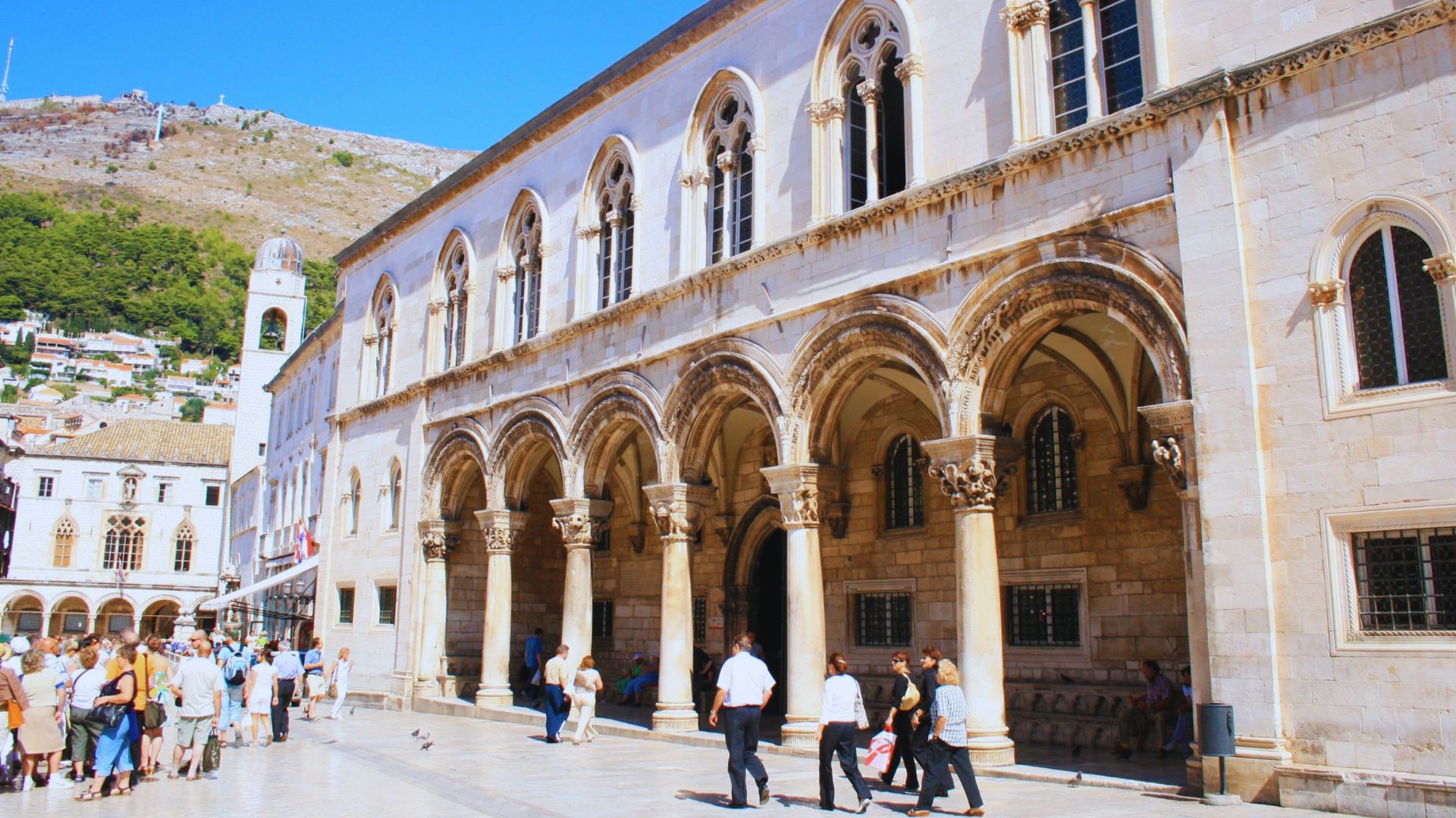 A historic building with arched colonnades and tall windows in a sunny European plaza, surrounded by tourists, with a hill in the background.