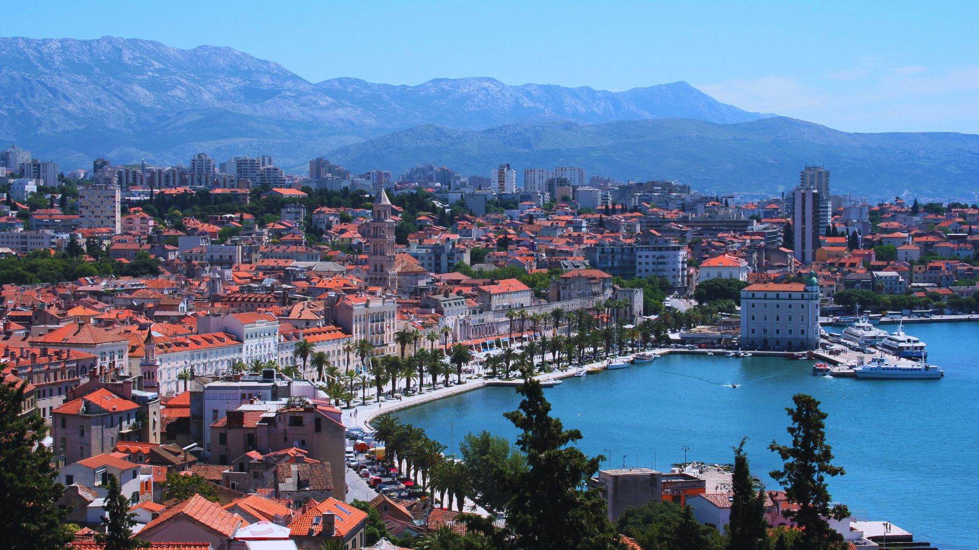 Aerial view of Split, Croatia, showcasing a scenic coastline with terracotta-roofed buildings, a bustling harbor, palm-lined promenade, and distant mountains.