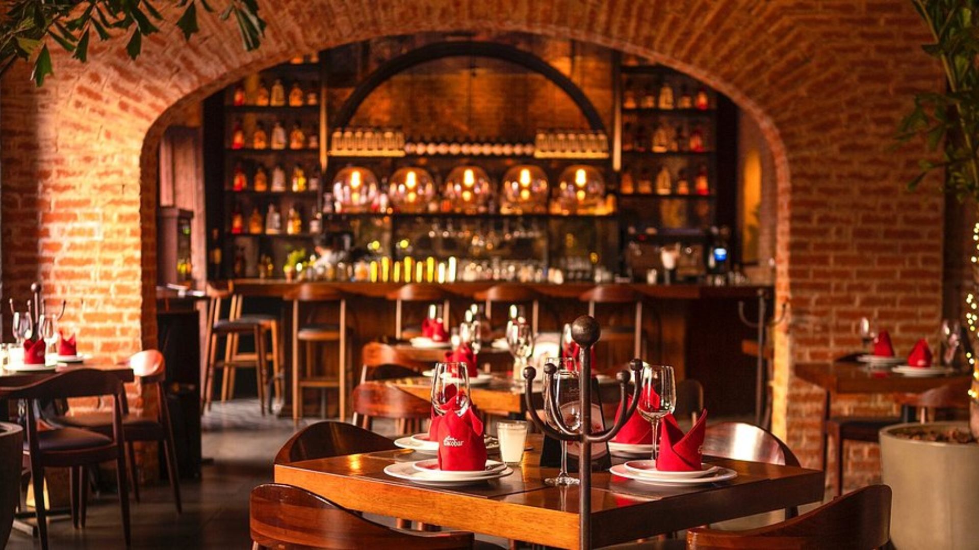 Cozy restaurant interior with brick arches, dim lighting, and wooden tables set with red napkins and glasses. Warm, inviting atmosphere with a well-stocked bar in the background.