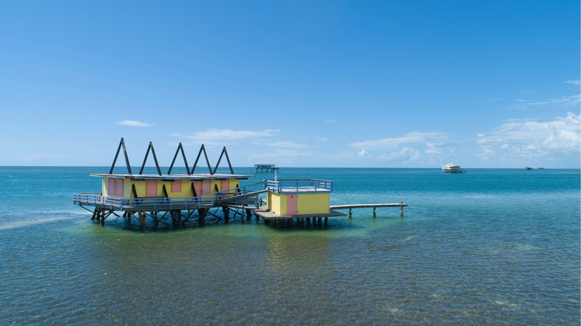 Colorful yellow and pink buildings on stilts above clear turquoise water. Blue sky with a few clouds, evoking a serene, tropical vibe.