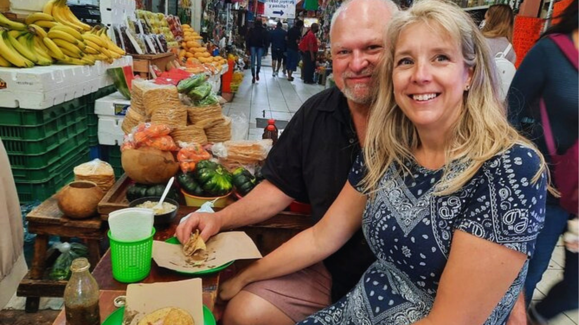 A man and woman seated at a table in a bustling market, surrounded by various stalls and shoppers.