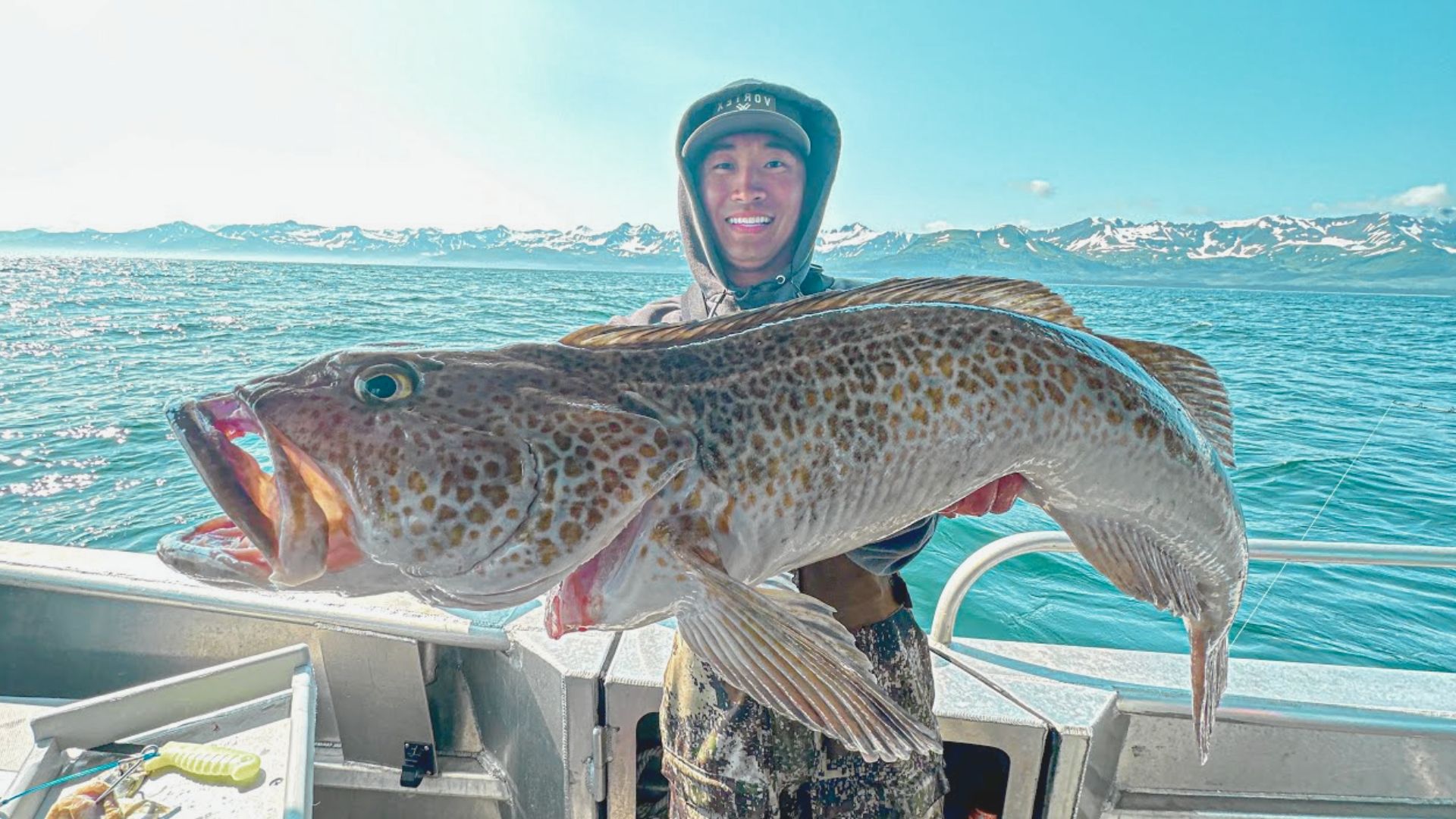 A person joyfully holds a large, speckled fish on a boat in clear, sunlight. The ocean and snow-capped mountains form a stunning backdrop.