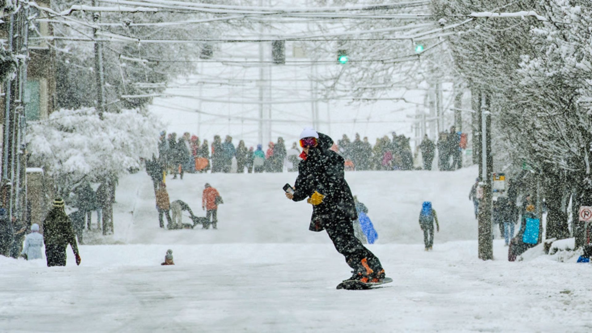 A person snowboards down a snowy city street lined with people, trees covered in snow, creating a playful, lively winter scene.
