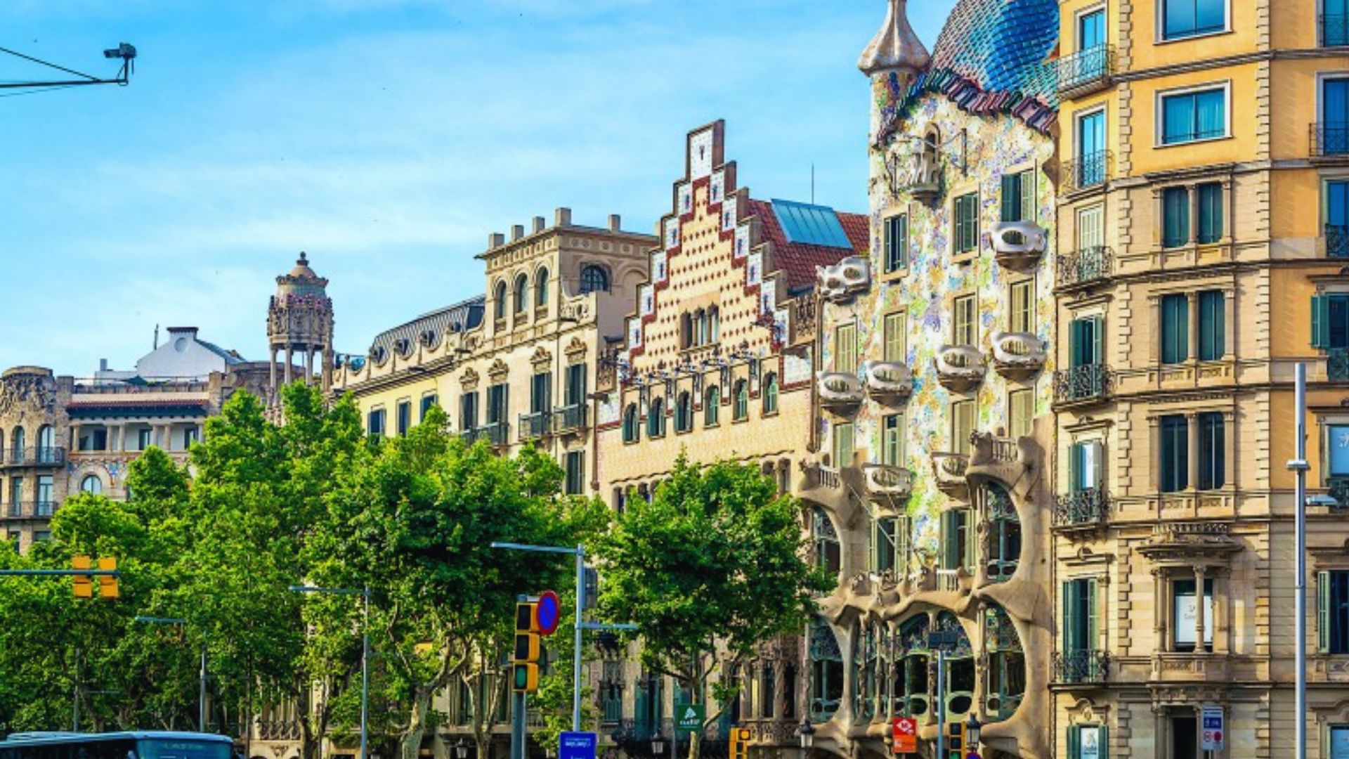 Street view showcasing colorful, ornate buildings with unique architecture, vibrant mosaic patterns, and lush green trees, under a clear blue sky.