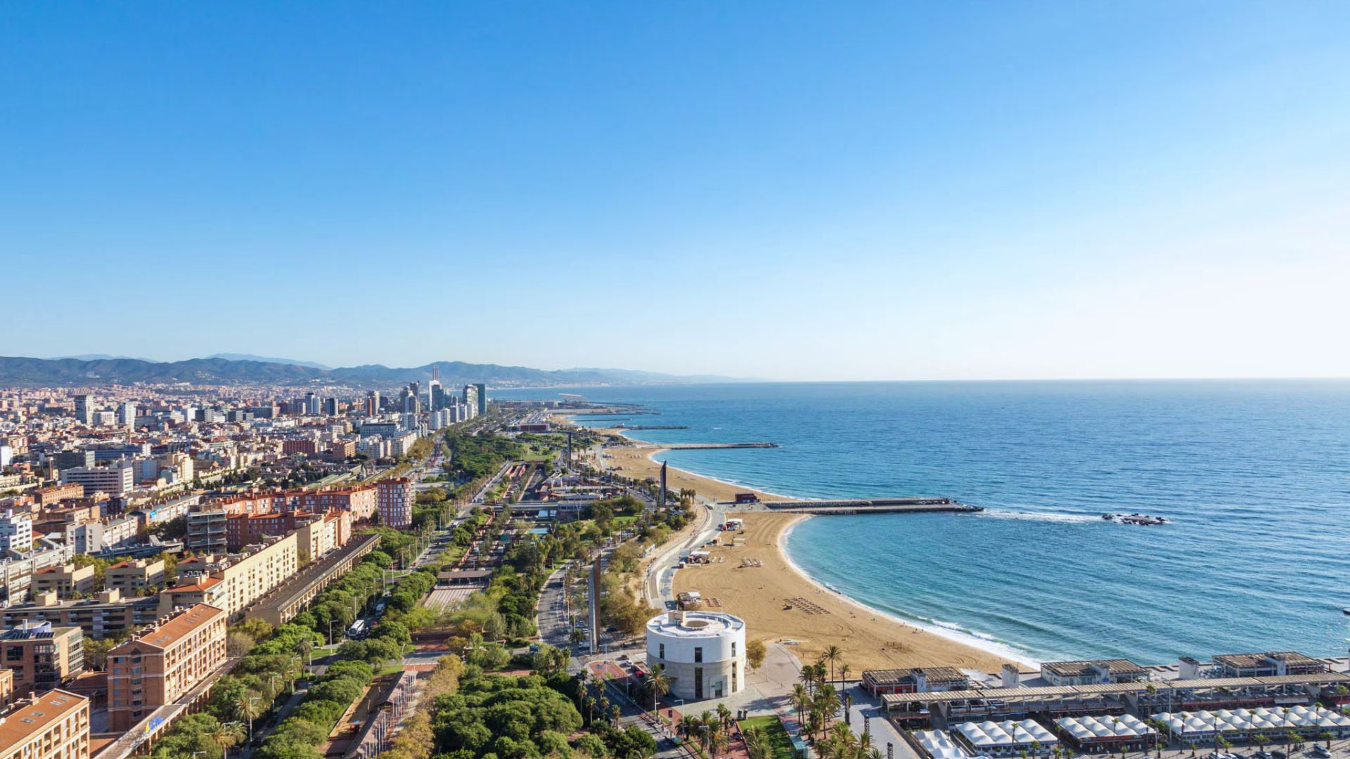 Aerial view of a coastal cityscape on a clear day. The image shows a sandy beach lining the blue sea, with urban buildings and green parks adjacent.