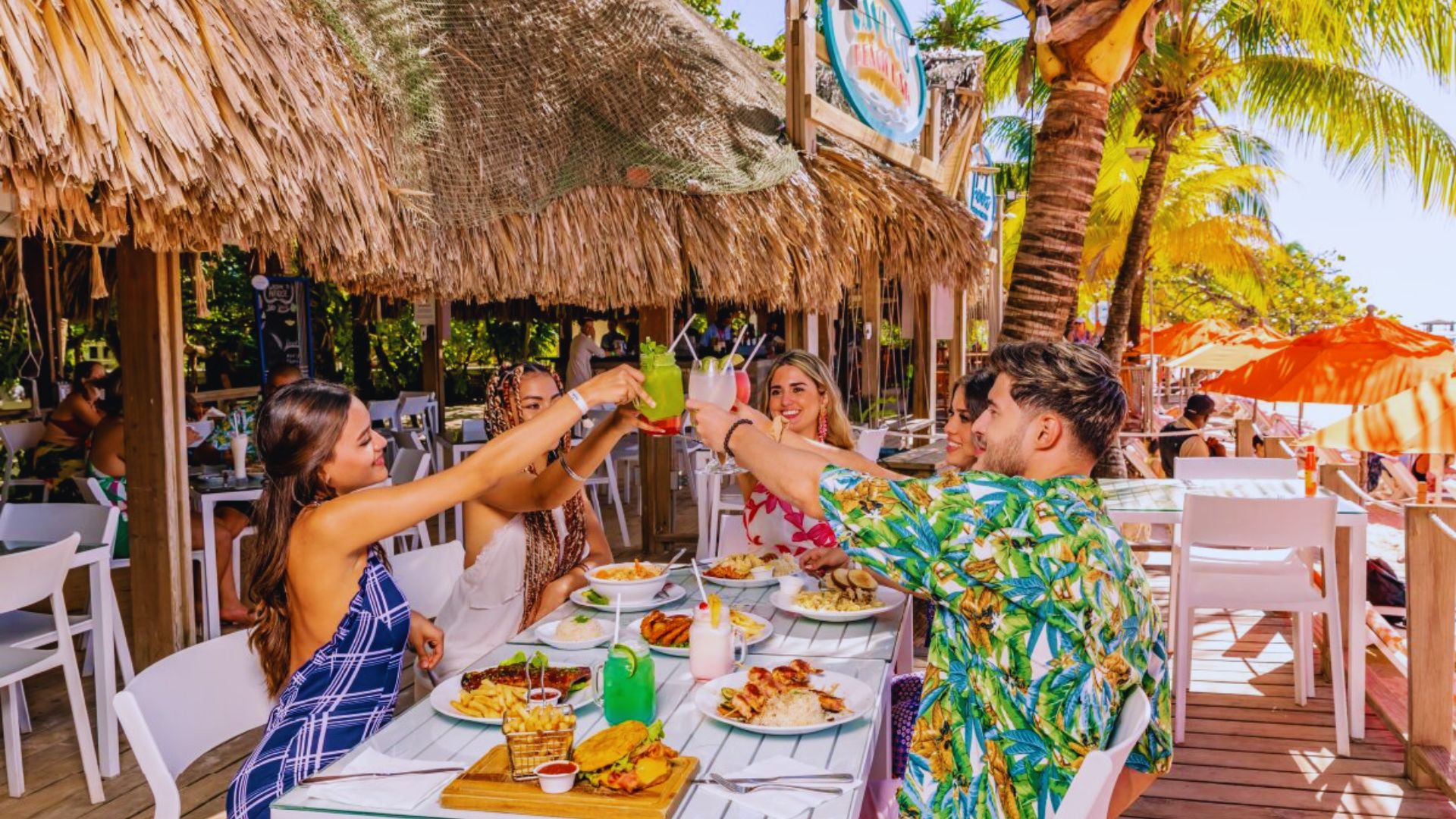Four friends toast with drinks at a tropical beachside restaurant. The setting features bright umbrellas, palm trees, and a wooden deck, creating a lively, vacation-like atmosphere.