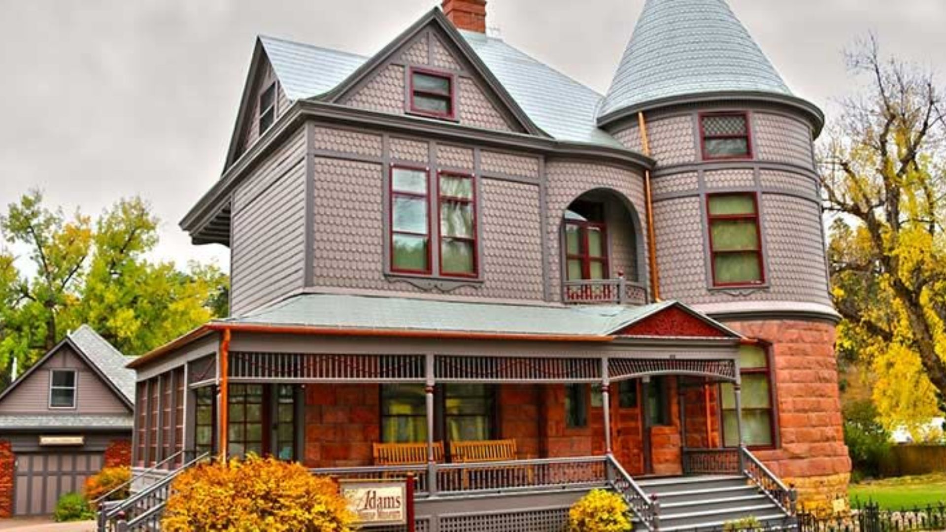A Victorian-style house with a turret and wraparound porch. The facade features intricate woodwork. Autumn trees and bushes surround the home.