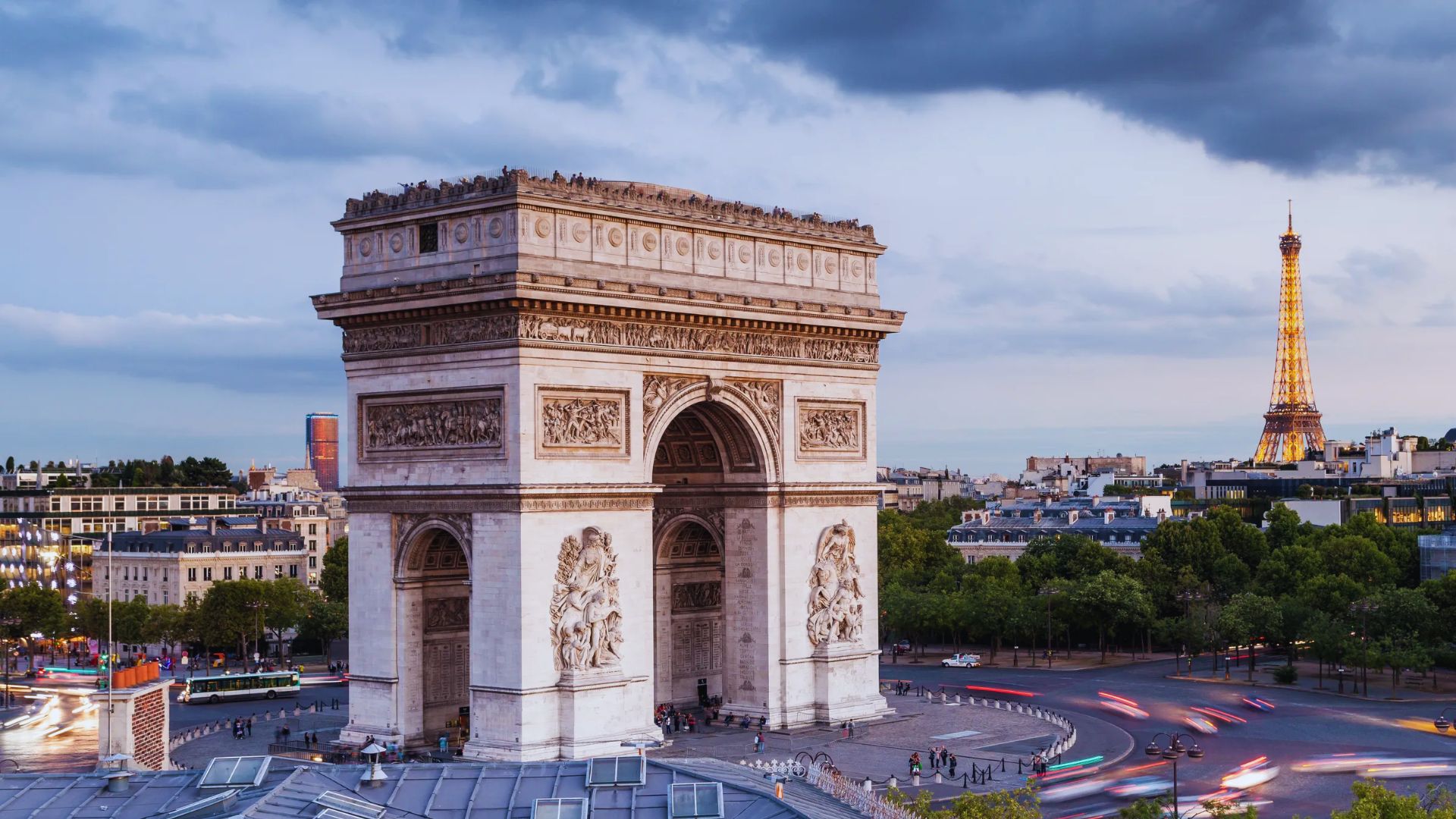 The image shows the Arc de Triomphe in Paris at dusk, with cars circling the monument. The Eiffel Tower is visible in the distance, under a cloudy sky.