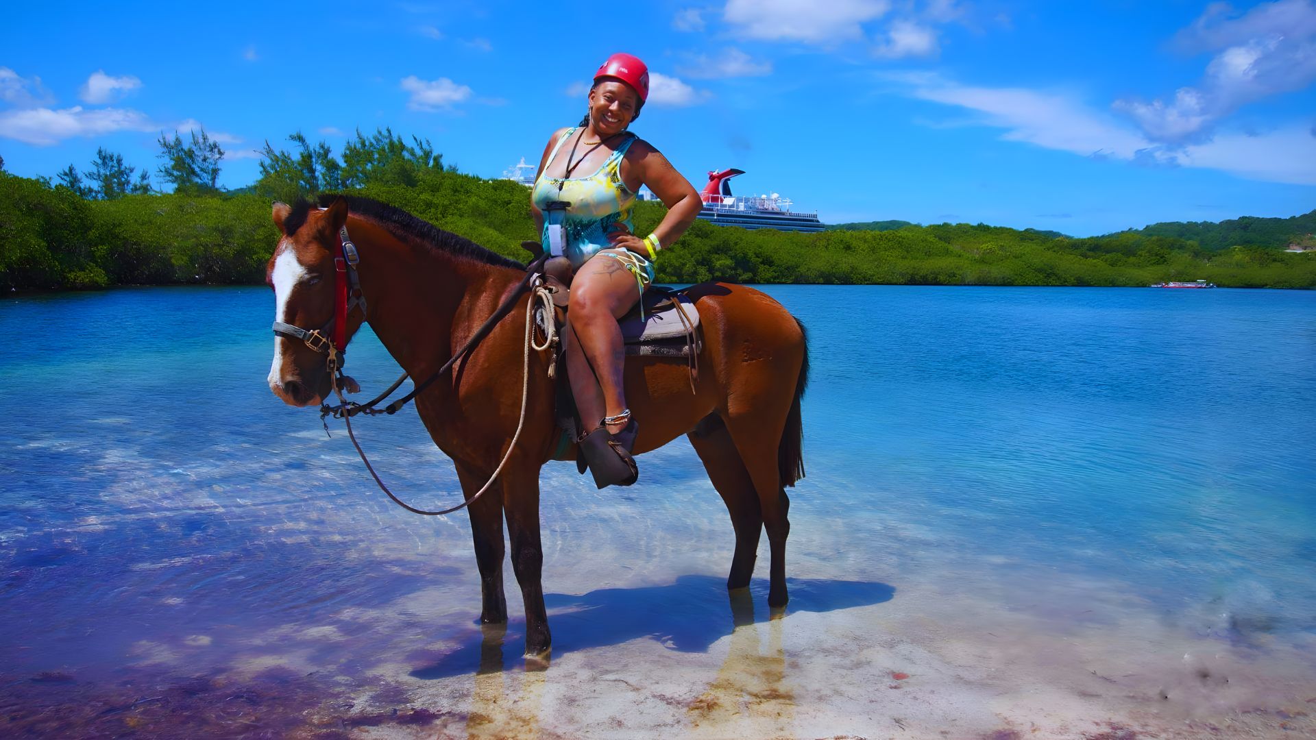 A person wearing a helmet smiles while riding a brown horse in shallow water with a cruise ship in the distance under a clear blue sky.