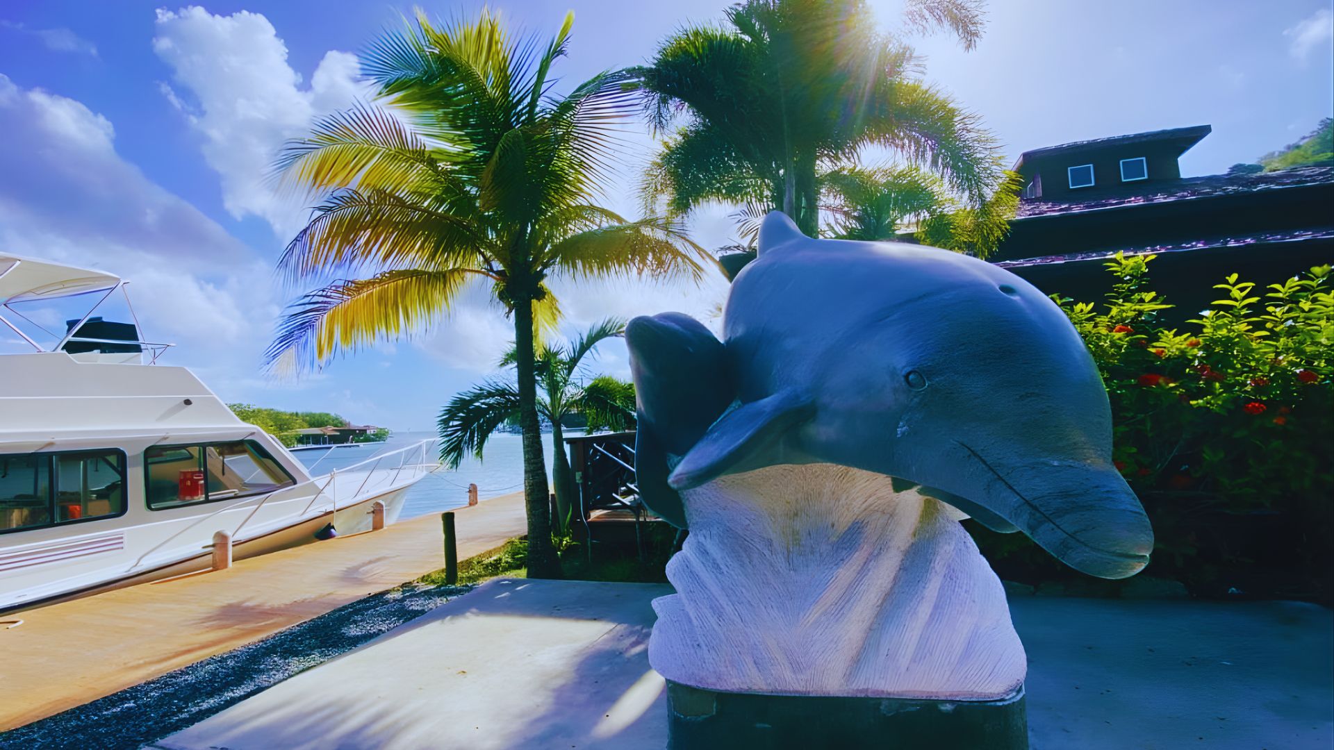 A dolphin statue stands on a sunny waterfront, flanked by palm trees. A white boat is docked nearby, with a clear blue sky and fluffy clouds above.