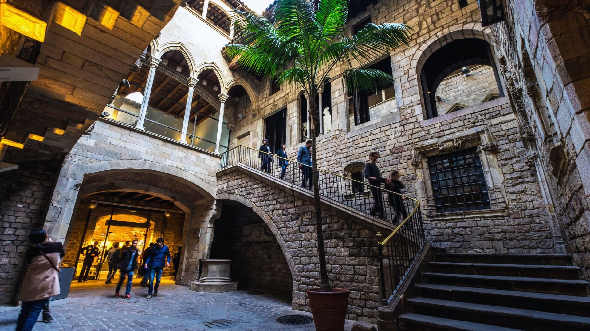Stone courtyard with arches, stairway, and a potted palm tree. People walk under warm lights, evoking a historic, peaceful atmosphere.