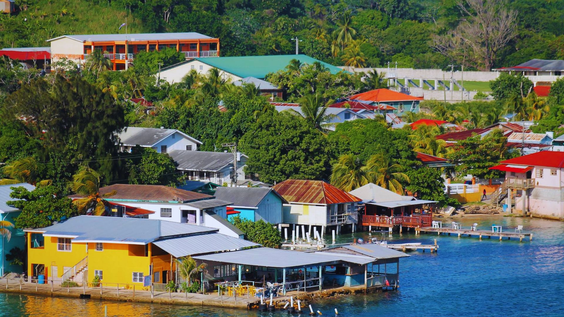 Colorful coastal village with vibrant houses, some on stilts, surrounded by lush greenery. The water is calm, conveying a serene and cheerful atmosphere.