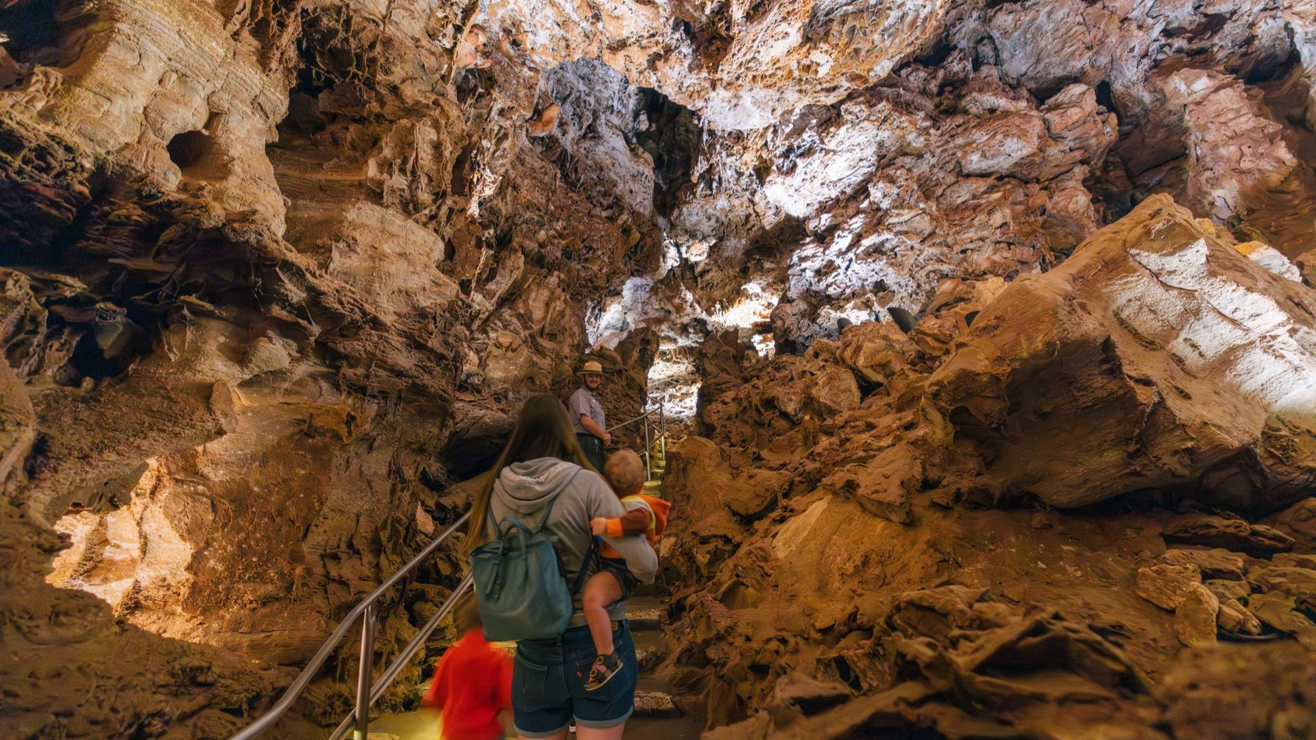 A family explores a dimly lit cave, with rough, textured rock walls. The mother carries a child while ascending stone steps, evoking a sense of adventure.