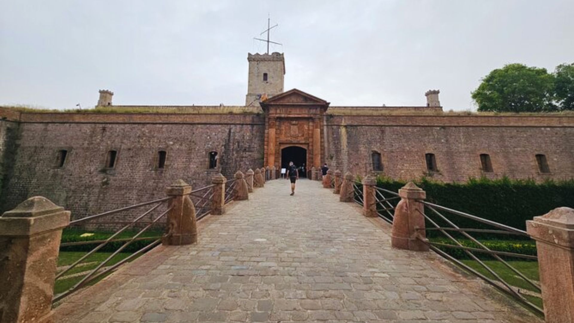 A cobblestone path leads to an ancient stone fortress with a central wooden door and tower. Overcast sky and sparse greenery create a historic atmosphere.