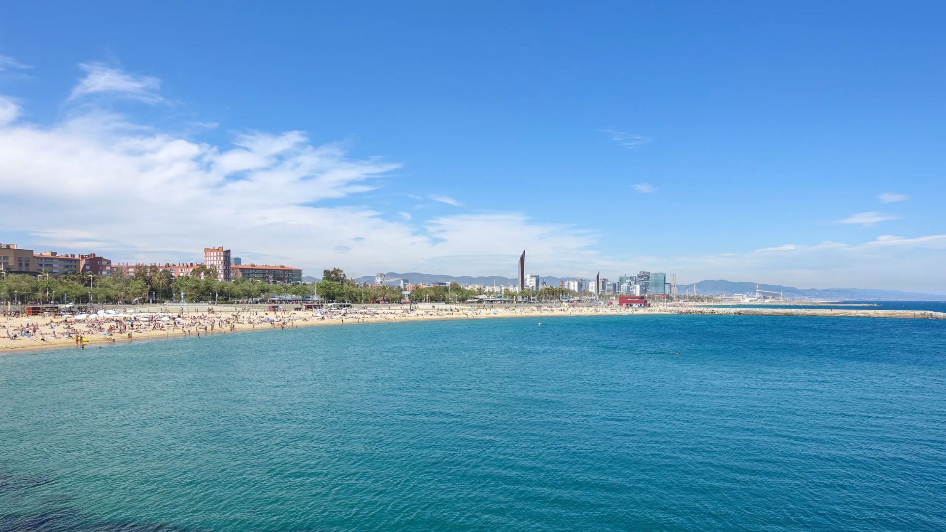 A sunny beach scene with clear blue water in the foreground and a crowded sandy beach in the middle. The city skyline and mountains are visible in the background.