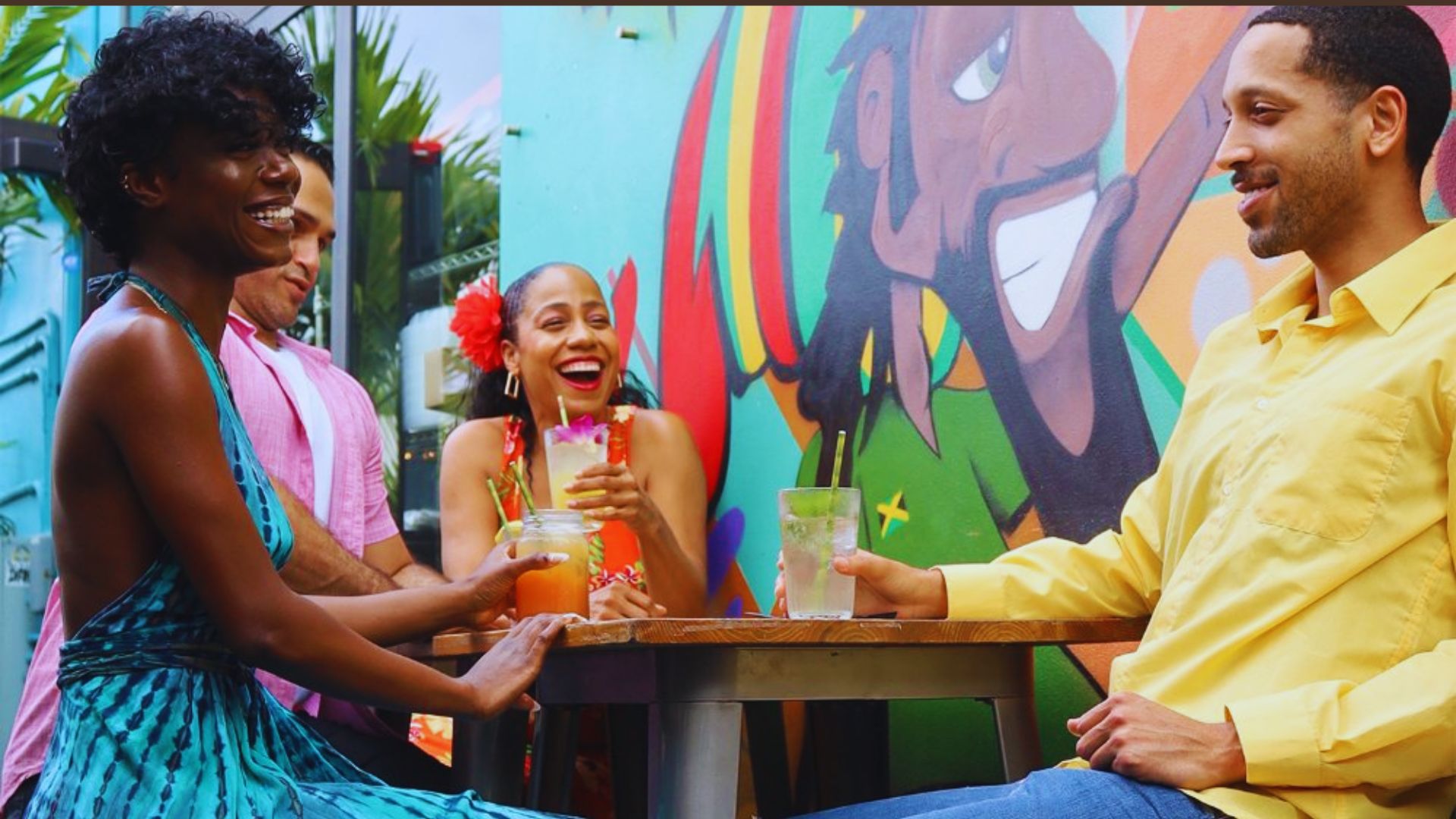 A group of four friends laughing and enjoying drinks at an outdoor table, set against a colorful mural. The scene is lively and joyful.