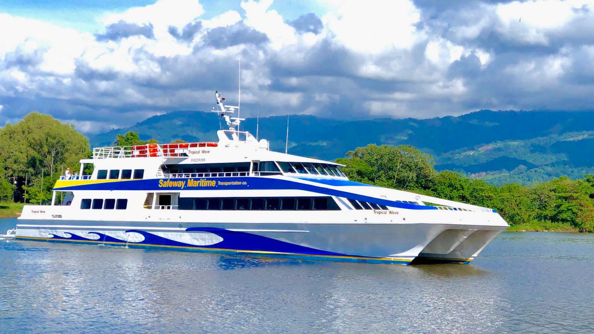 A sleek white and blue ferry with "Safeway Maritime" branding sails on a calm lake, set against a backdrop of lush green hills and a cloudy sky.