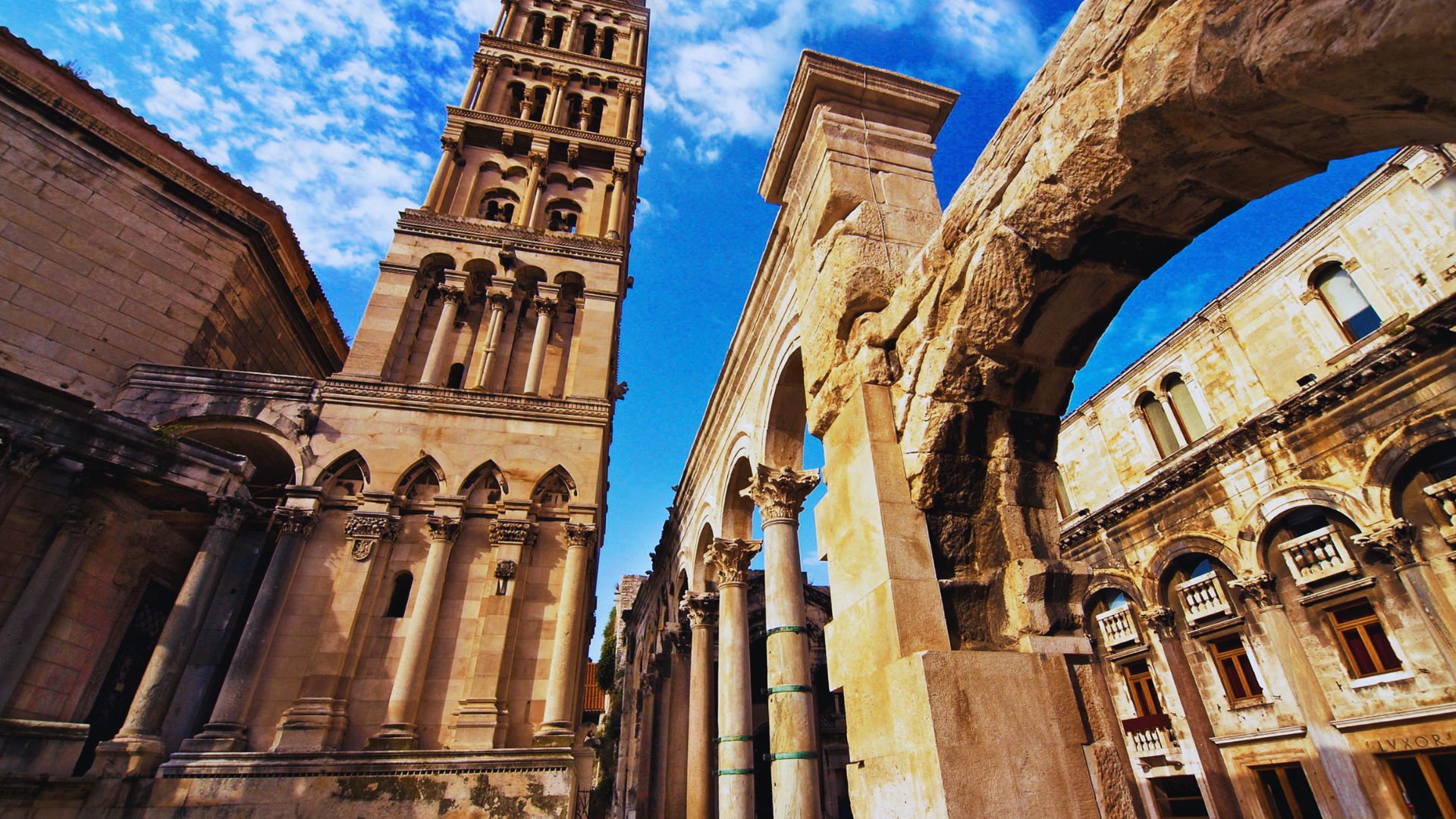Ancient stone archways and a tall, ornate bell tower under a vibrant blue sky with clouds; evokes a sense of historical grandeur and architectural beauty.