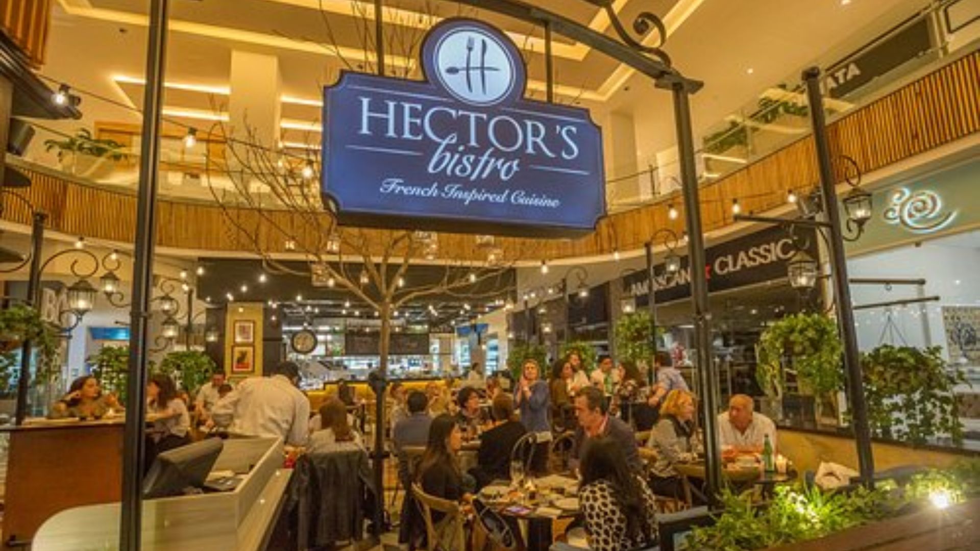 Bustling bistro scene at a shopping mall, featuring a sign for "Hector's Bistro." Diners enjoy French-inspired cuisine in a lively, warmly-lit setting.