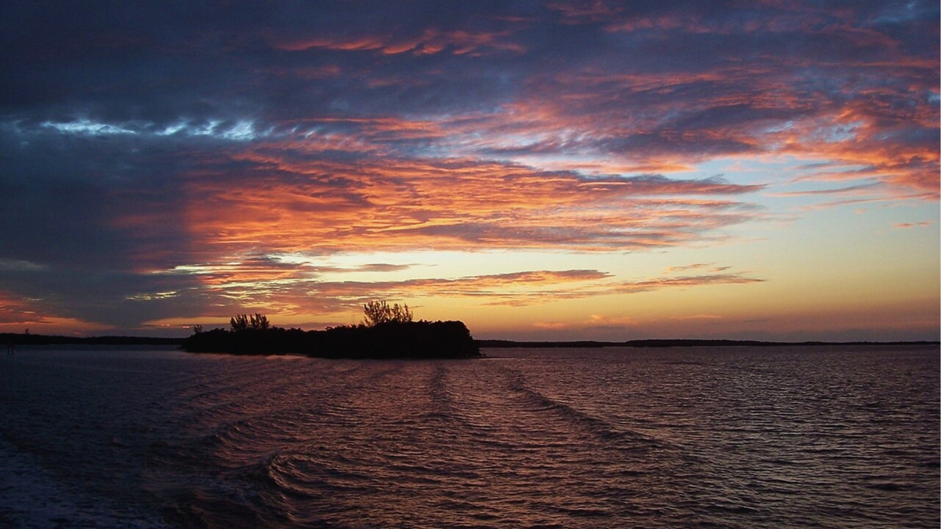 Sunset over a calm sea with vibrant orange, pink, and purple clouds. Silhouette of land on the horizon, creating a tranquil and serene atmosphere.
