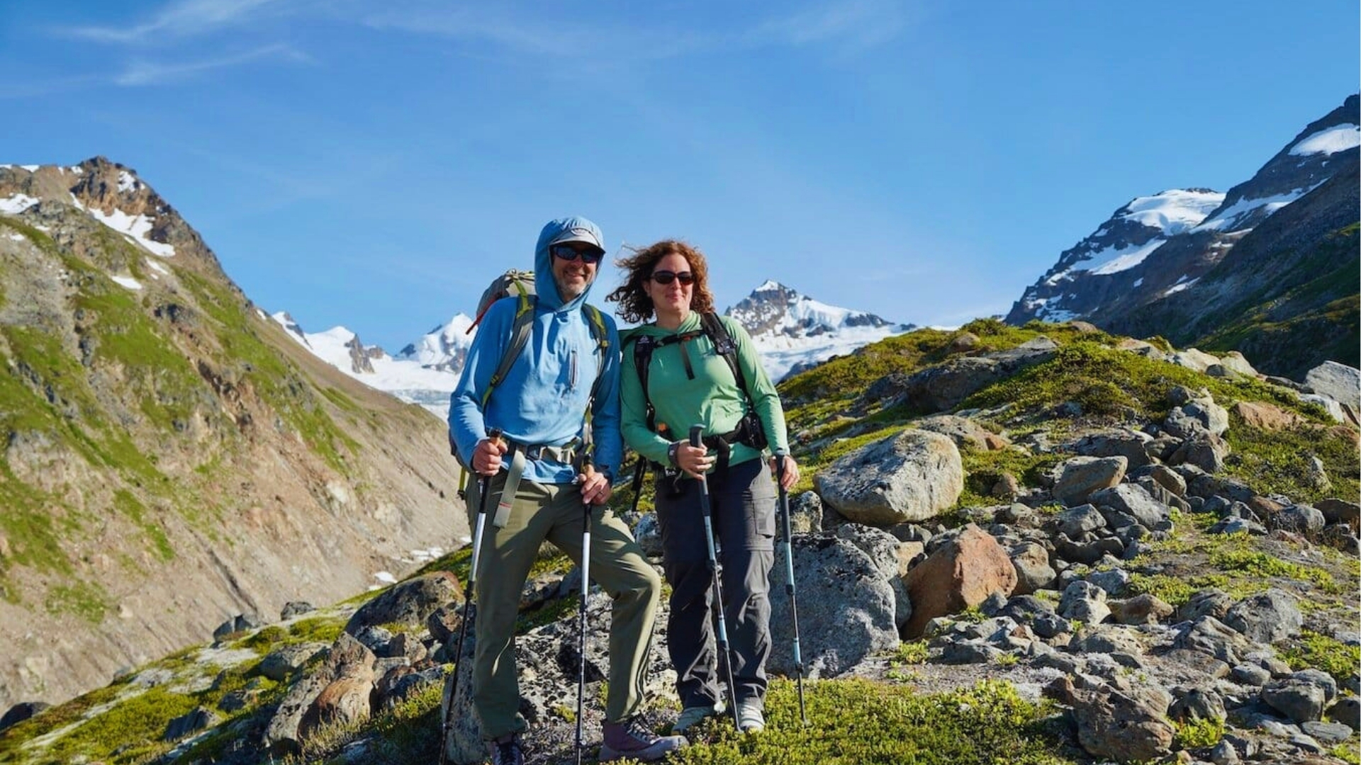 A couple hikes in a scenic mountain landscape, smiling and holding trekking poles. Snow-capped peaks and a clear blue sky create a serene backdrop.