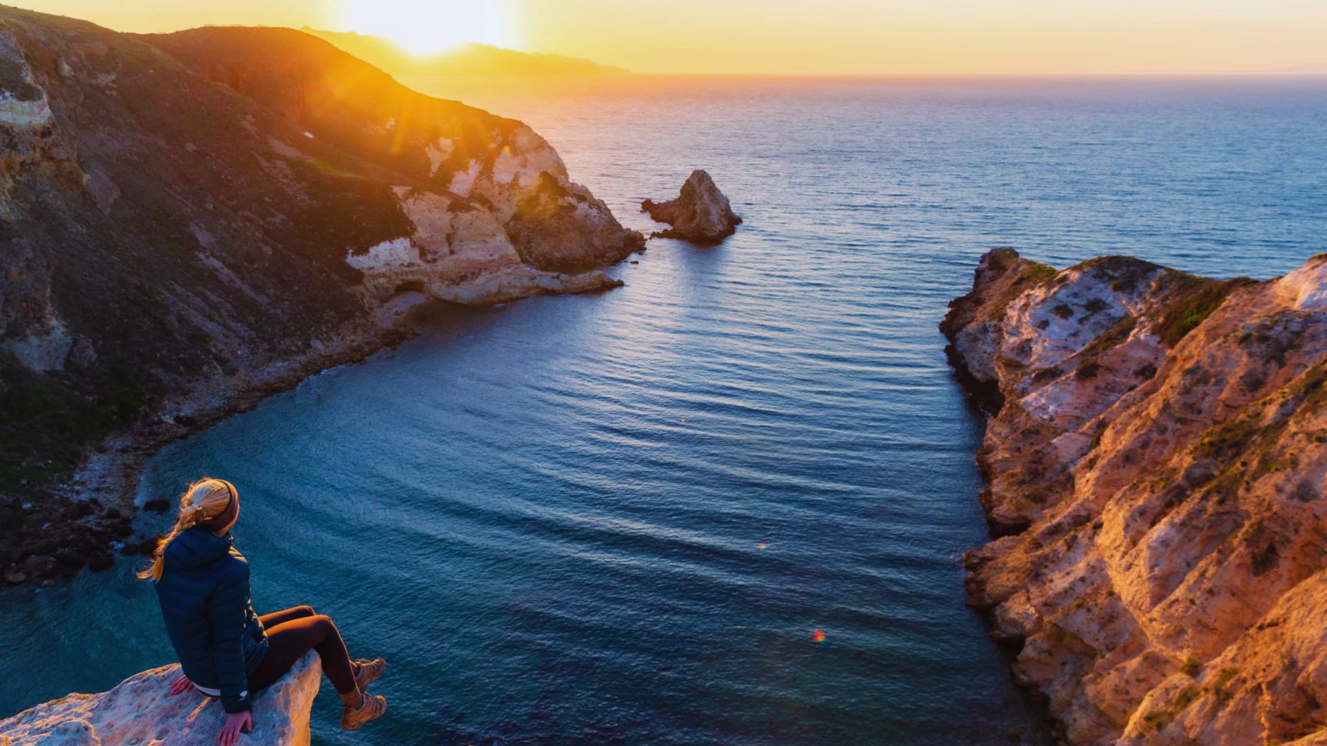 Woman sitting on a cliff's edge, gazing at the ocean during a vibrant sunset.