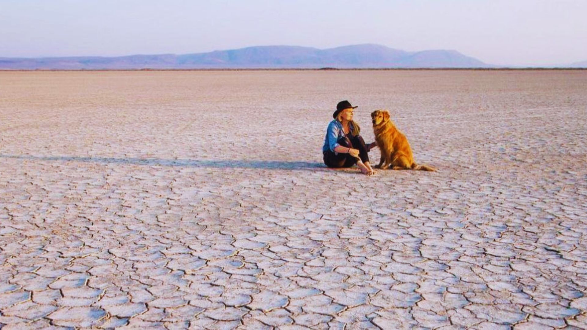 A man and his dog sit together in a vast desert landscape under a clear blue sky.