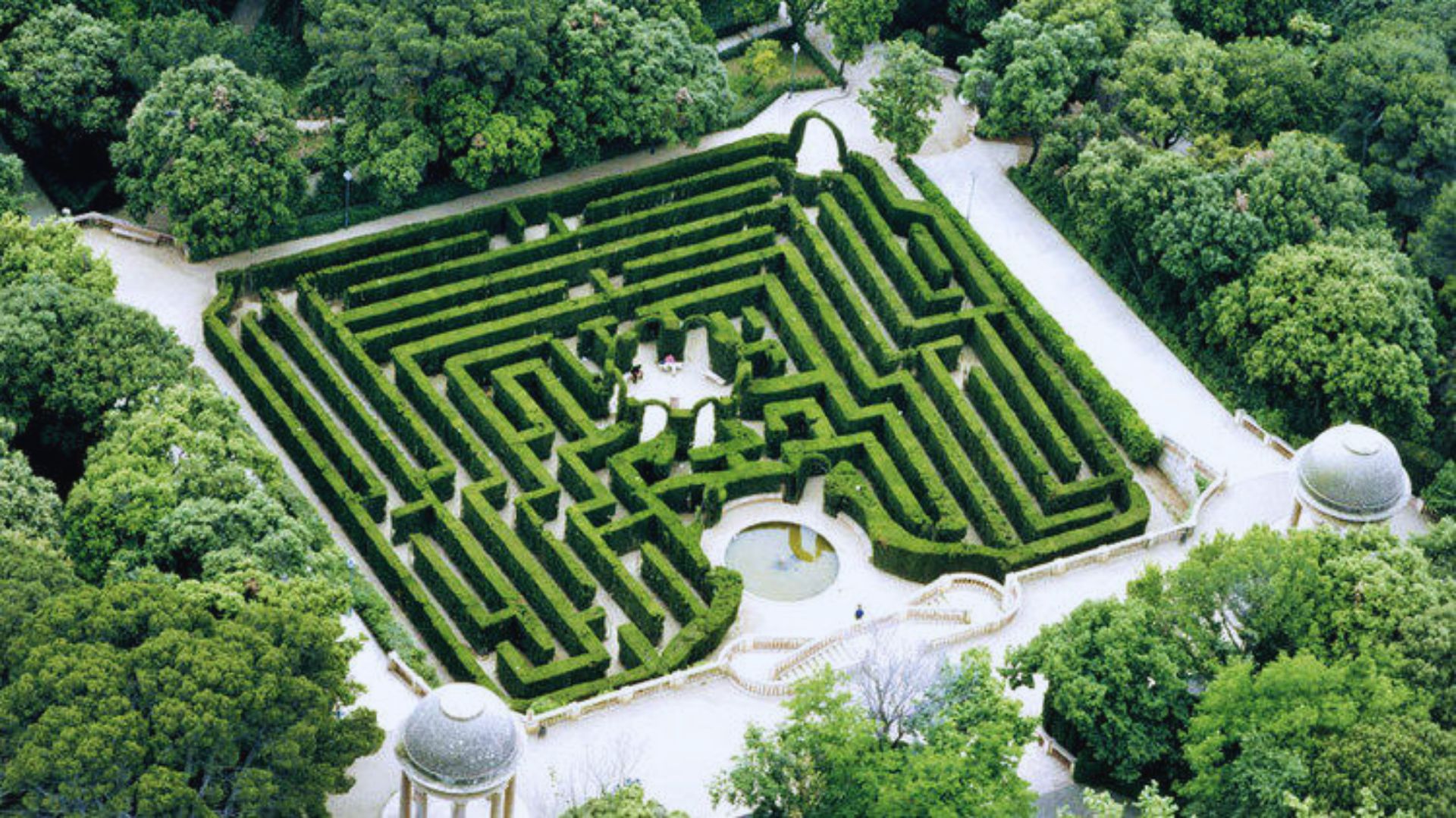 Aerial view of a lush, green hedge maze surrounded by trees. The maze features winding paths and a central circular area, creating a serene atmosphere.