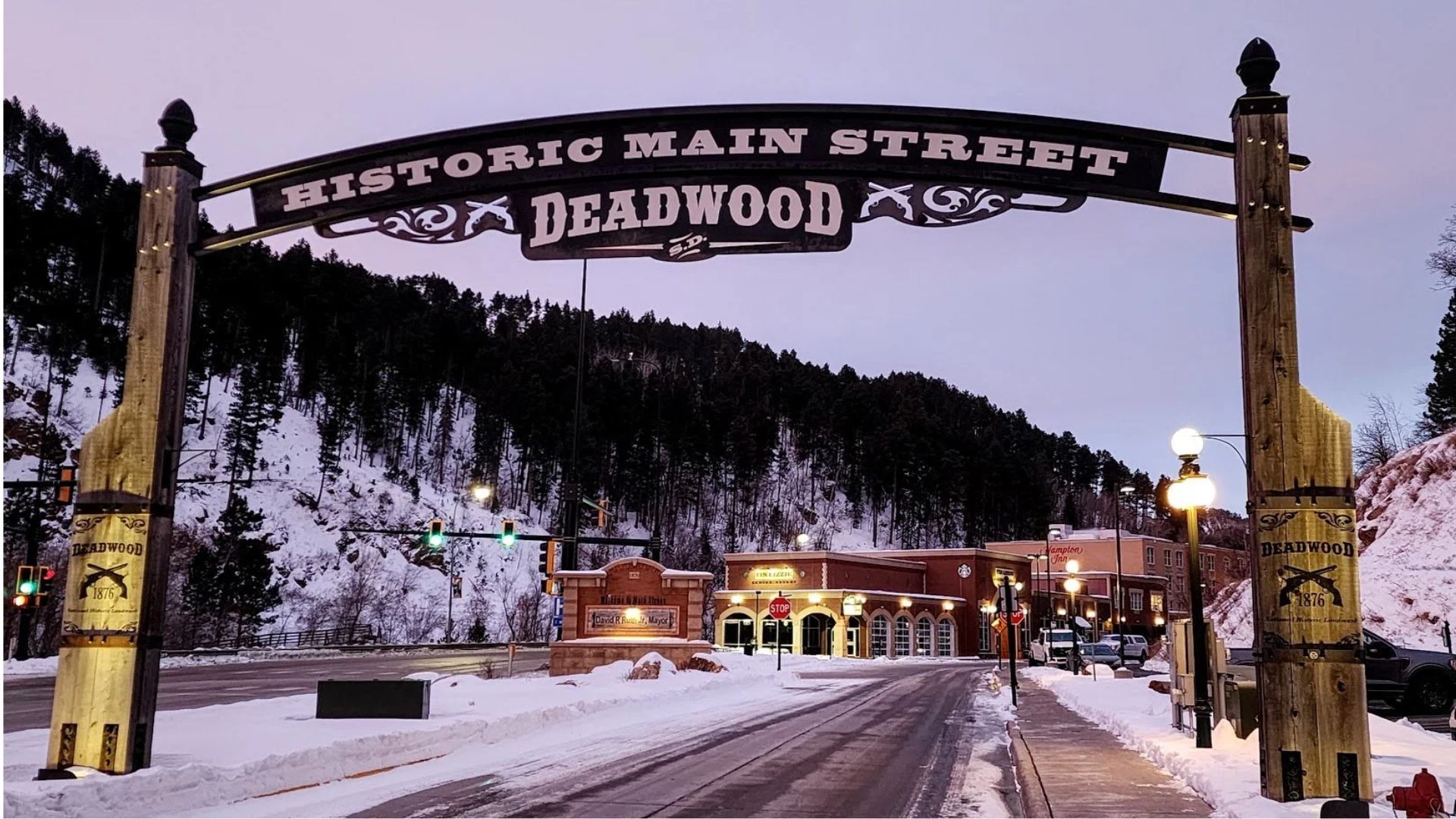 Archway reading "Historic Main Street Deadwood" over a snowy street, with scenic mountains and lit storefronts in the background, creating a historic, cozy atmosphere.