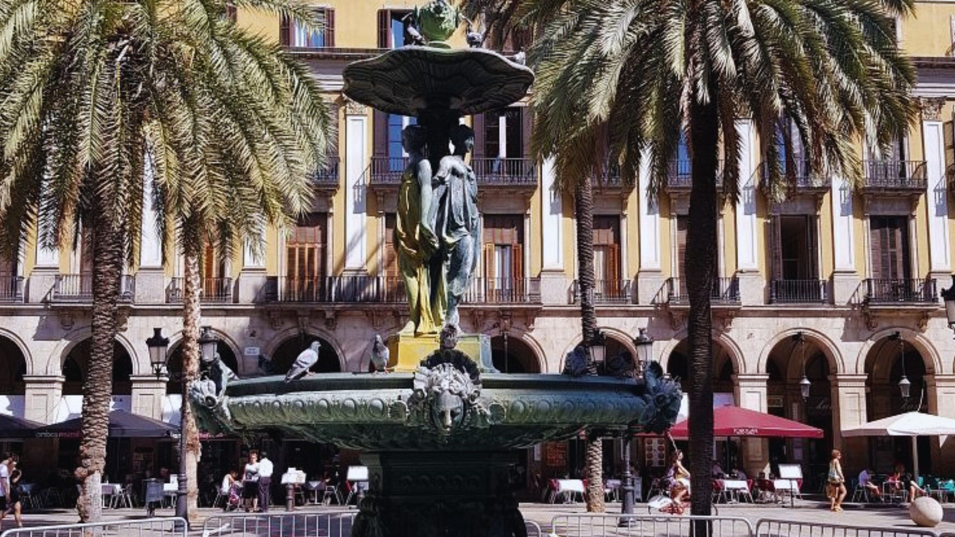 A historic fountain with ornate sculptures stands in a sunny plaza surrounded by palm trees and a yellow building with arched balconies, evoking a lively, warm atmosphere.