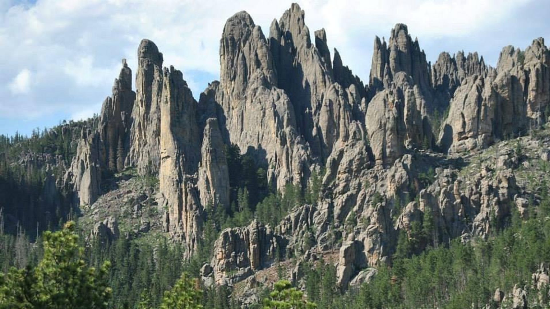 Jagged rock formations rise dramatically against a backdrop of blue sky and scattered clouds, with lush green pine trees at their base.