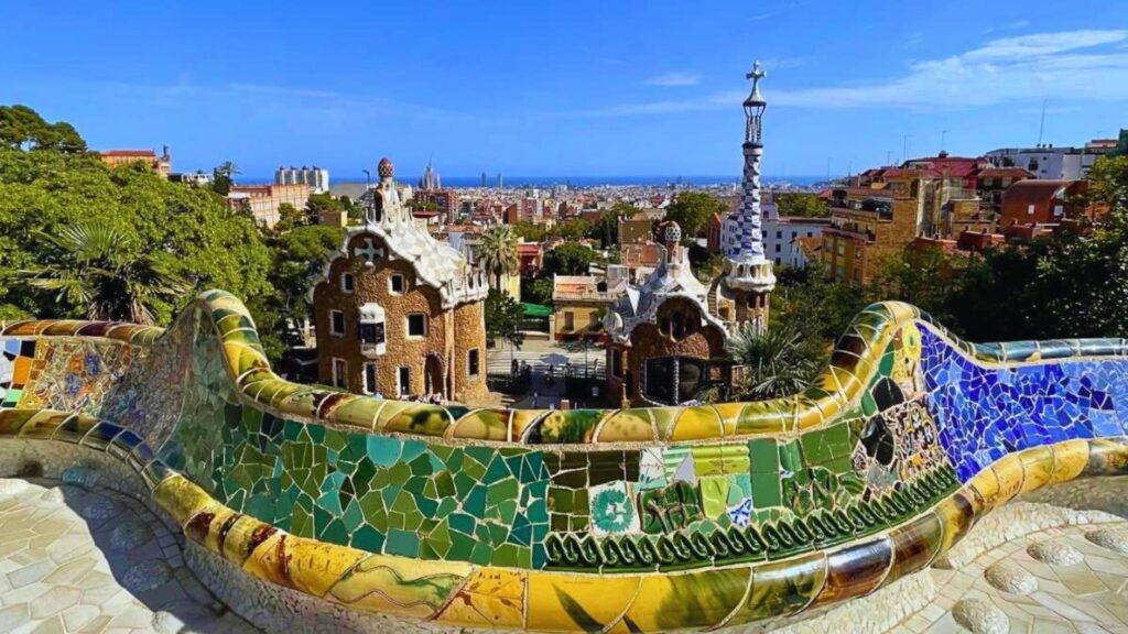 Colorful mosaic bench overlooks whimsical buildings and lush greenery in Park Güell, Barcelona, under a vibrant blue sky. Urban skyline in the distance.