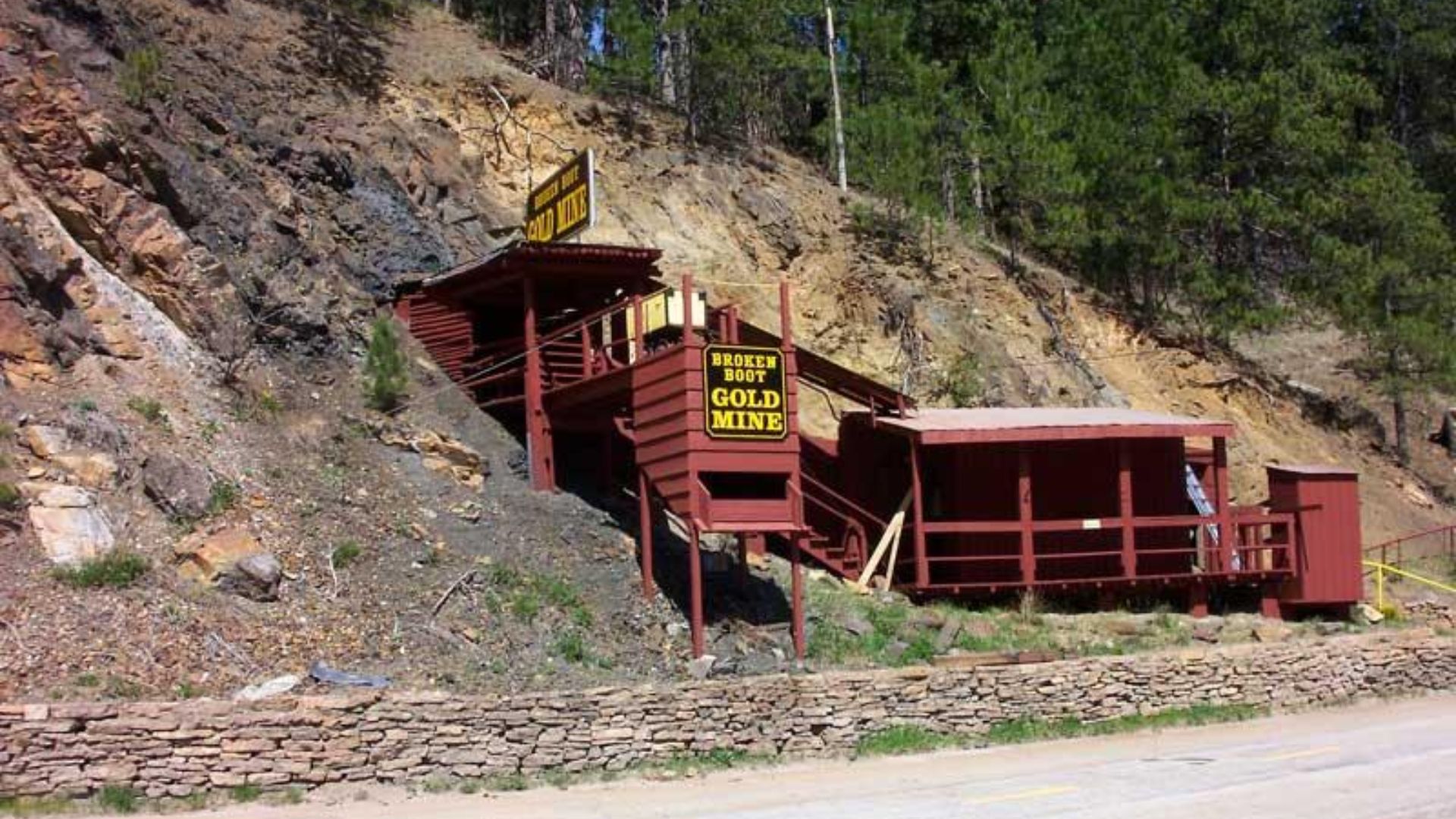 Rustic wooden structures of the Broken Boot Gold Mine are built into a rocky hillside. Signs with yellow letters indicate the entrance. Forested area above.