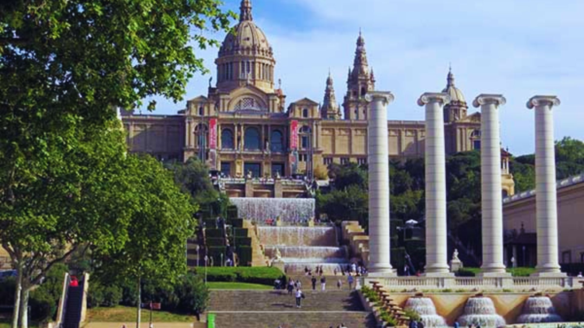 Grand building with a dome and spires, set behind cascading fountains and tall columns, surrounded by lush greenery under a clear blue sky.