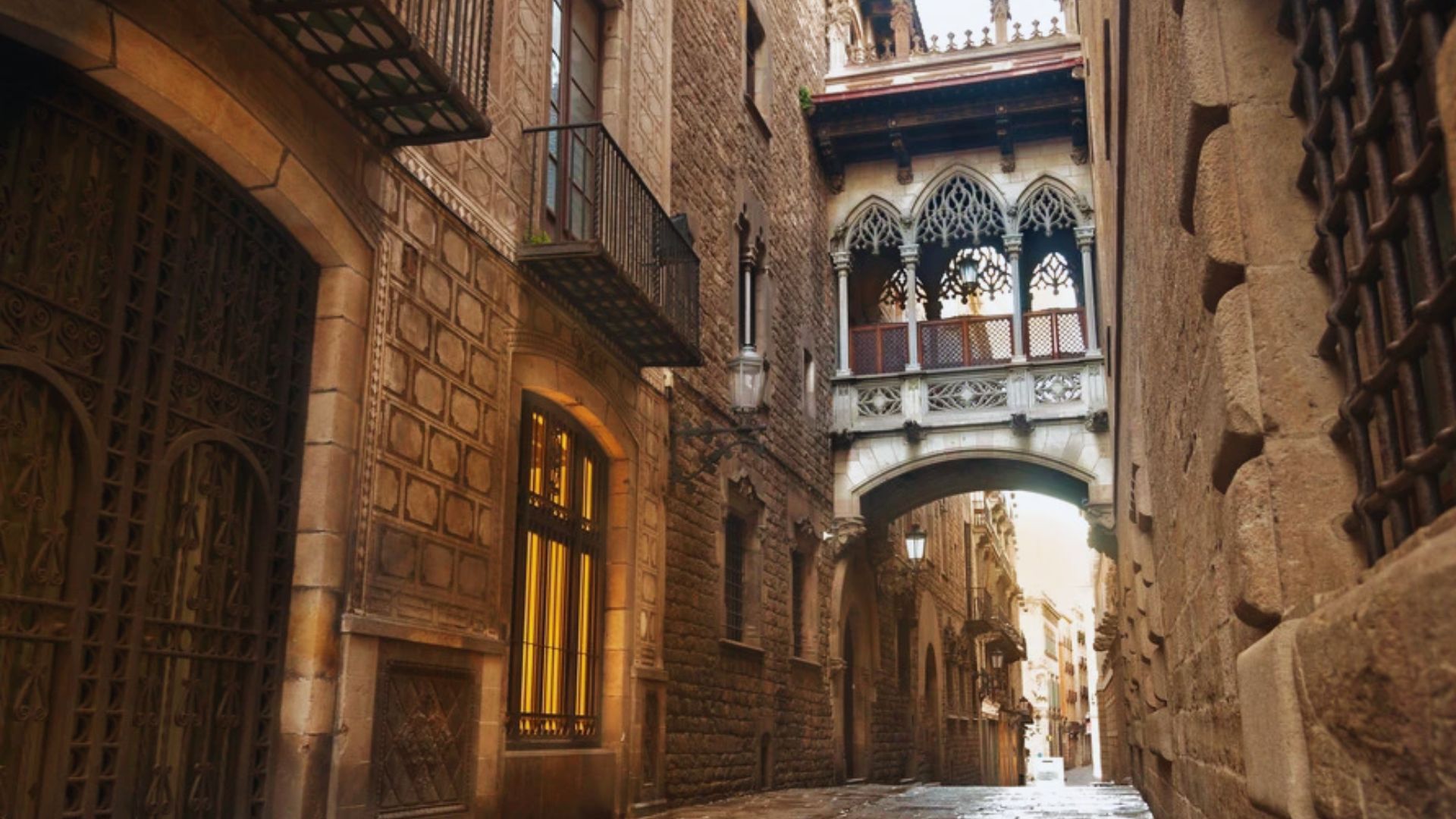 Narrow cobblestone alley in a Gothic Quarter, lined with ornate medieval buildings. An intricately designed bridge arches overhead, adding historical charm.