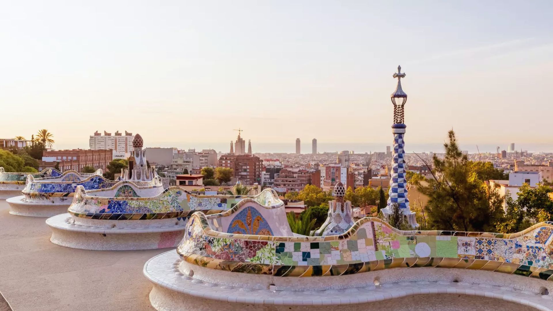 Panoramic view of Park Güell, Barcelona, at sunset. Vibrant mosaic benches curve in the foreground with a cityscape and sky in soft hues behind.