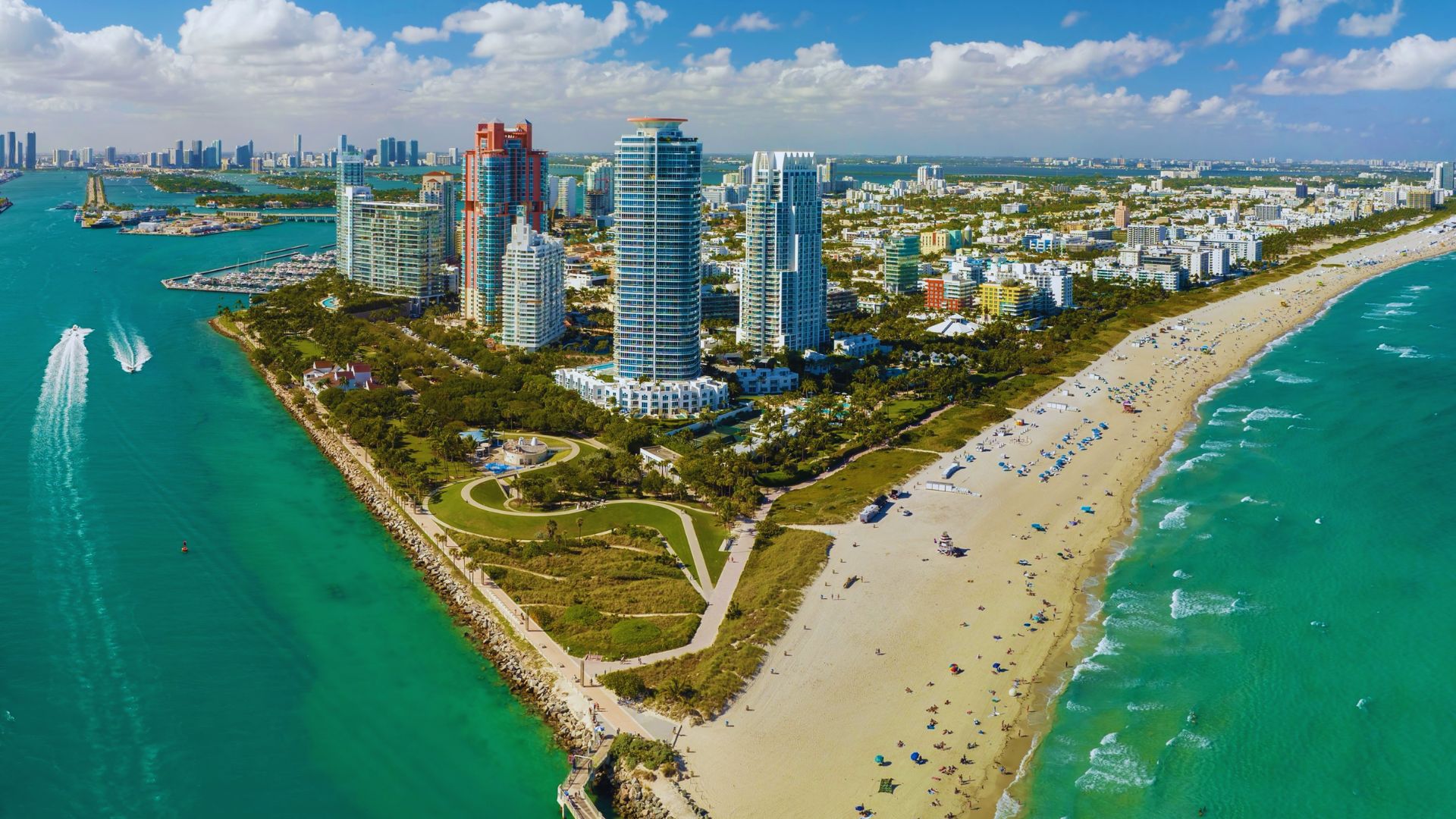 Aerial view of a vibrant Miami coastline with turquoise waters, sandy beaches, and lush greenery. Skyscrapers tower in the background under a blue sky.