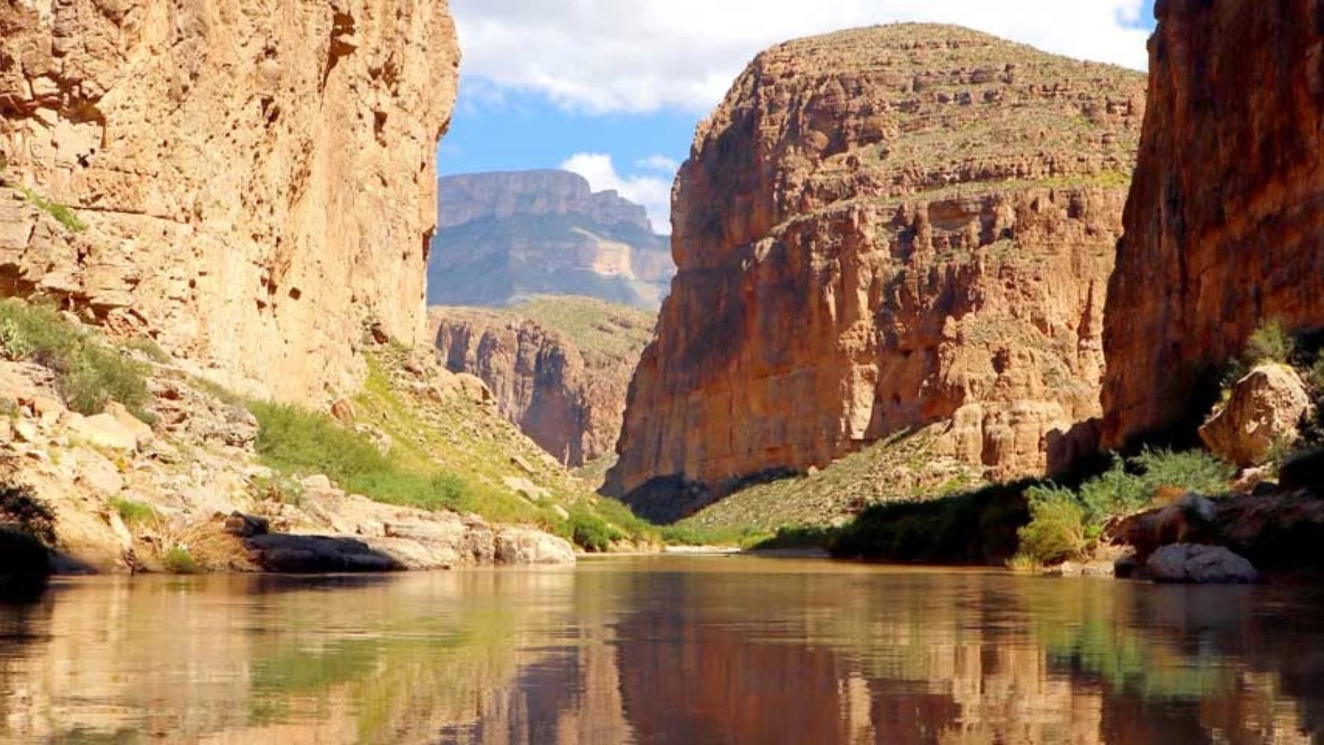 A serene river winds through a rocky canyon with steep, sunlit cliffs. Sparse greenery adorns the cliffs, and the calm water reflects the vibrant blue sky.