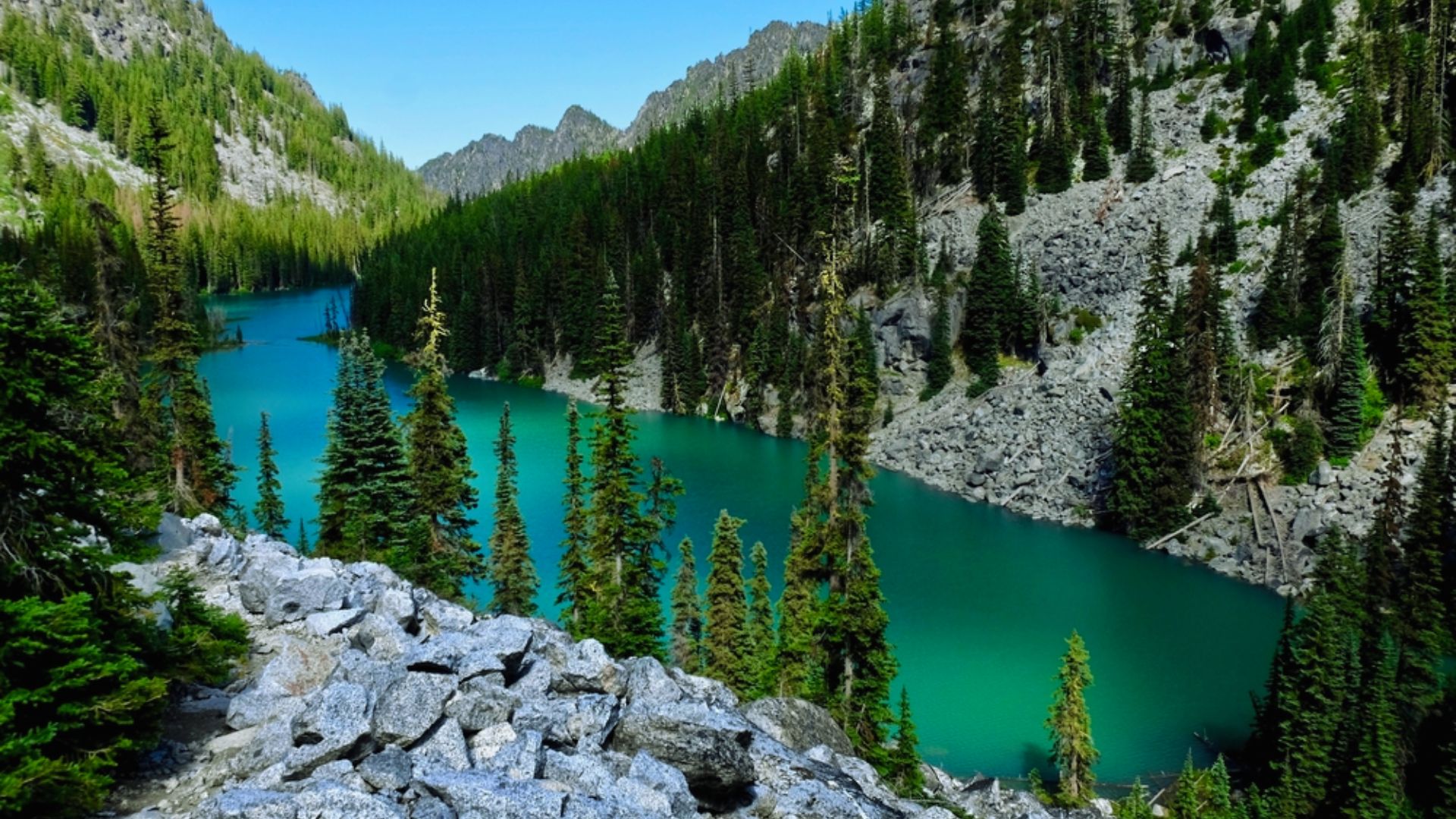A serene lake bordered by lush trees and majestic mountains under a clear blue sky.