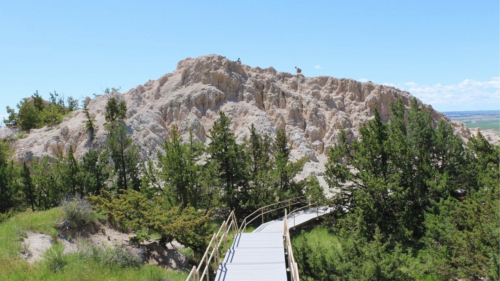 A wooden walkway extends towards a rocky outcropping, surrounded by natural scenery.