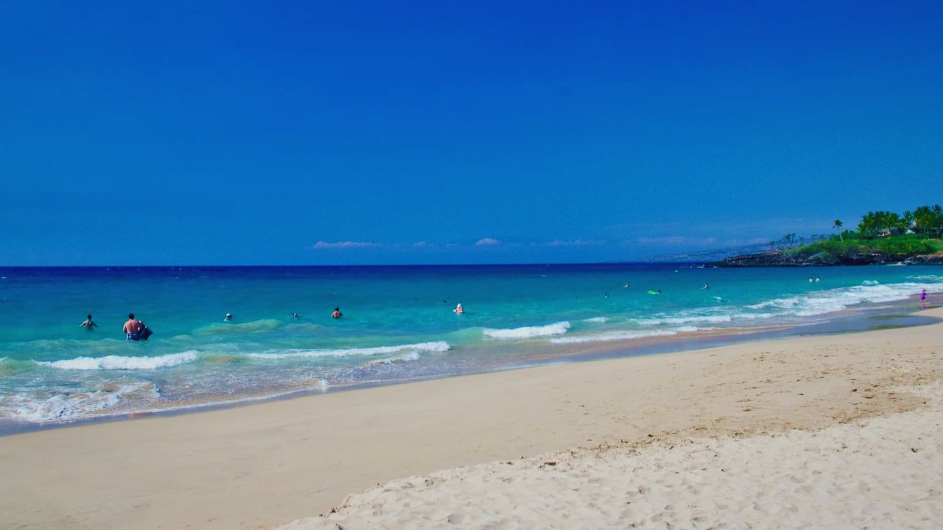 A sunny beach scene with several people swimming in the clear blue water, enjoying a warm day by the shore.