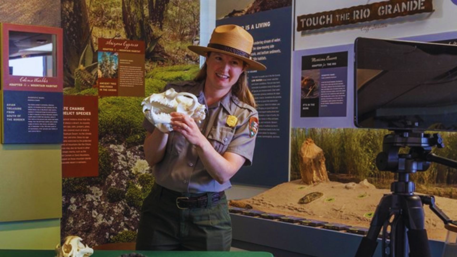 A woman wearing a hat holds a stuffed animal, smiling gently at the camera in a bright outdoor setting.