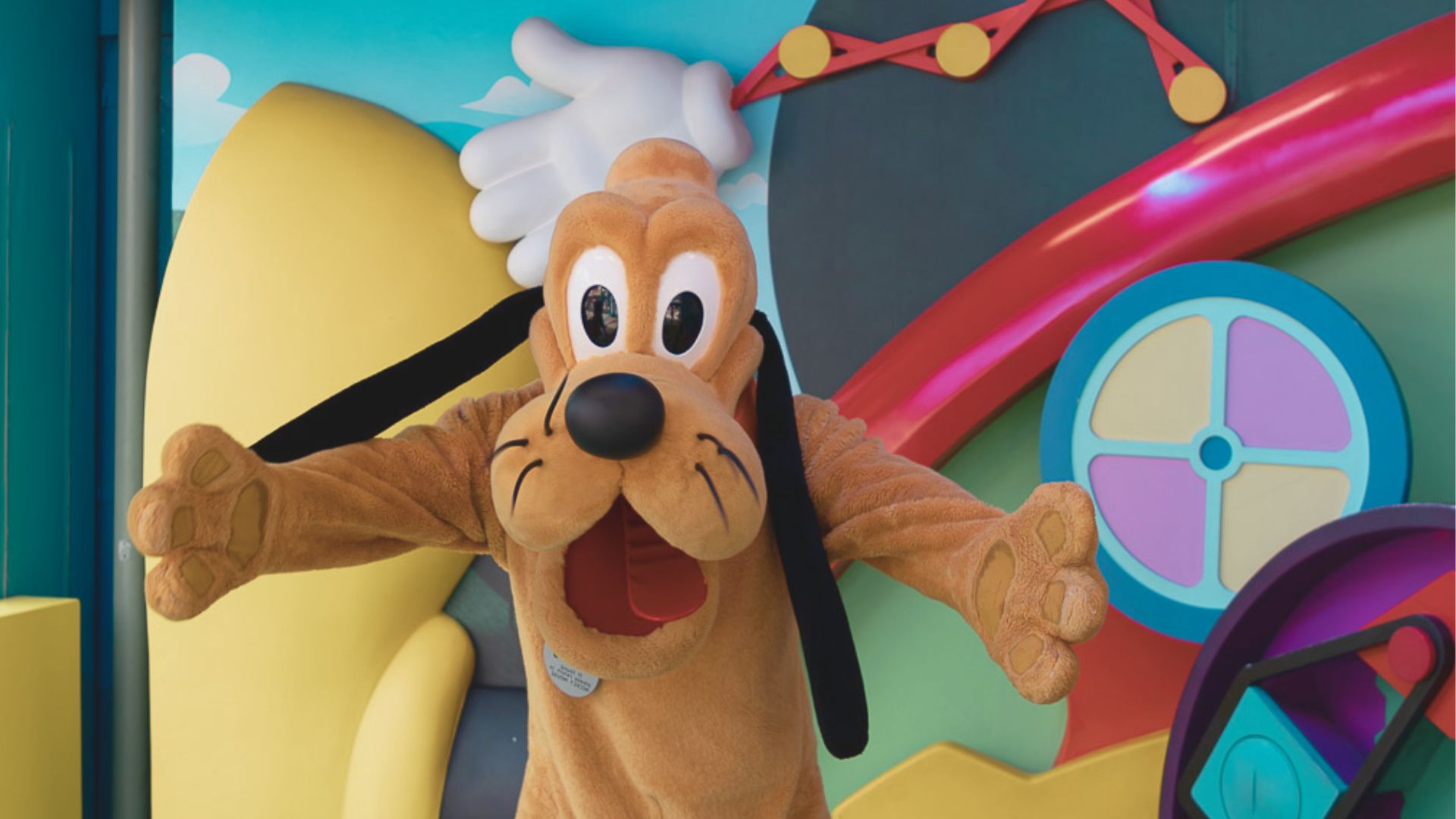 A cartoon character in a brown dog costume is joyfully waving in a colorful, whimsical setting with bright shapes and playful designs in the background.