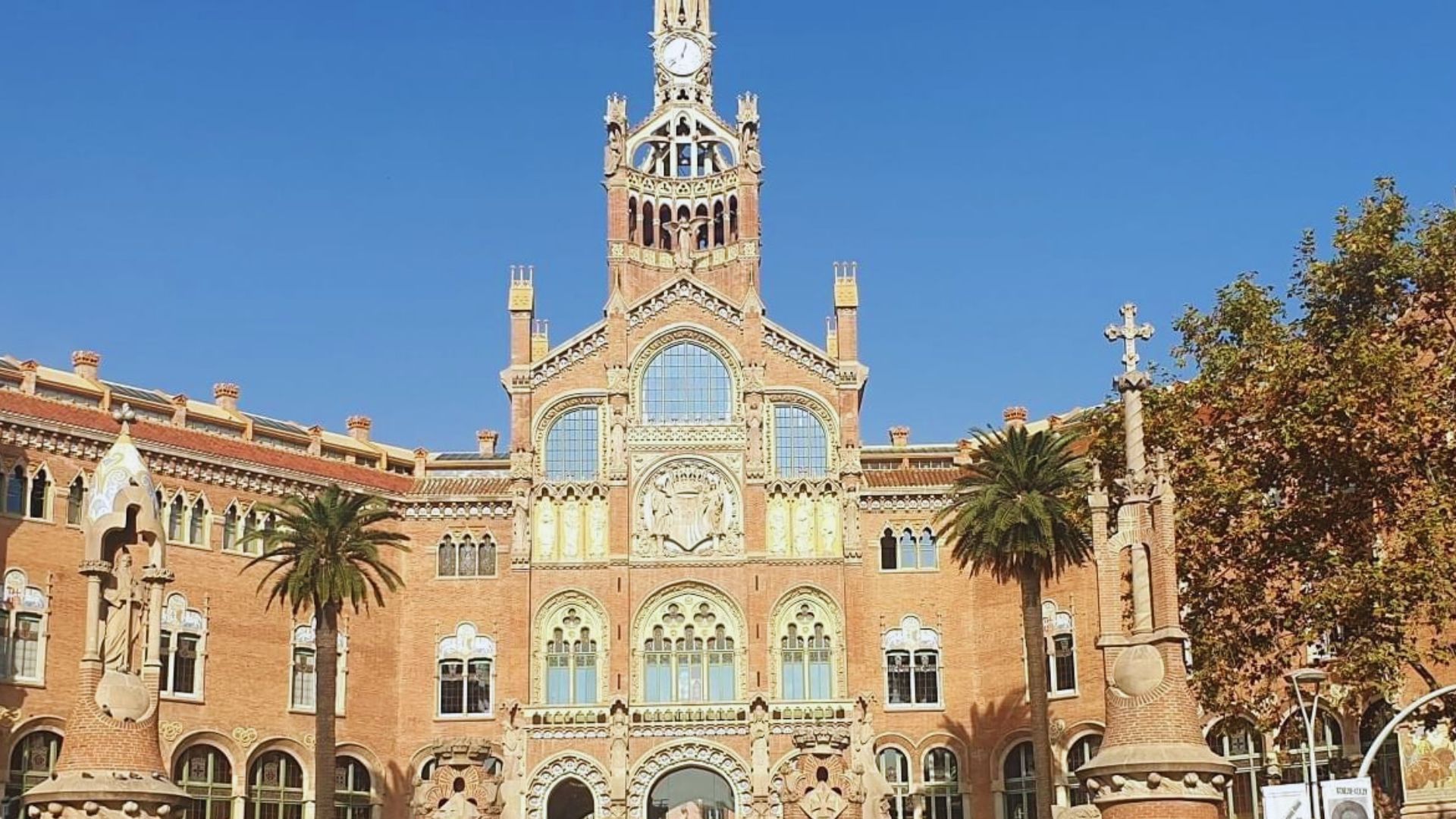 A stunning historic building with intricate brickwork and arched windows stands tall under a clear blue sky, flanked by palm trees and ornate sculptures.